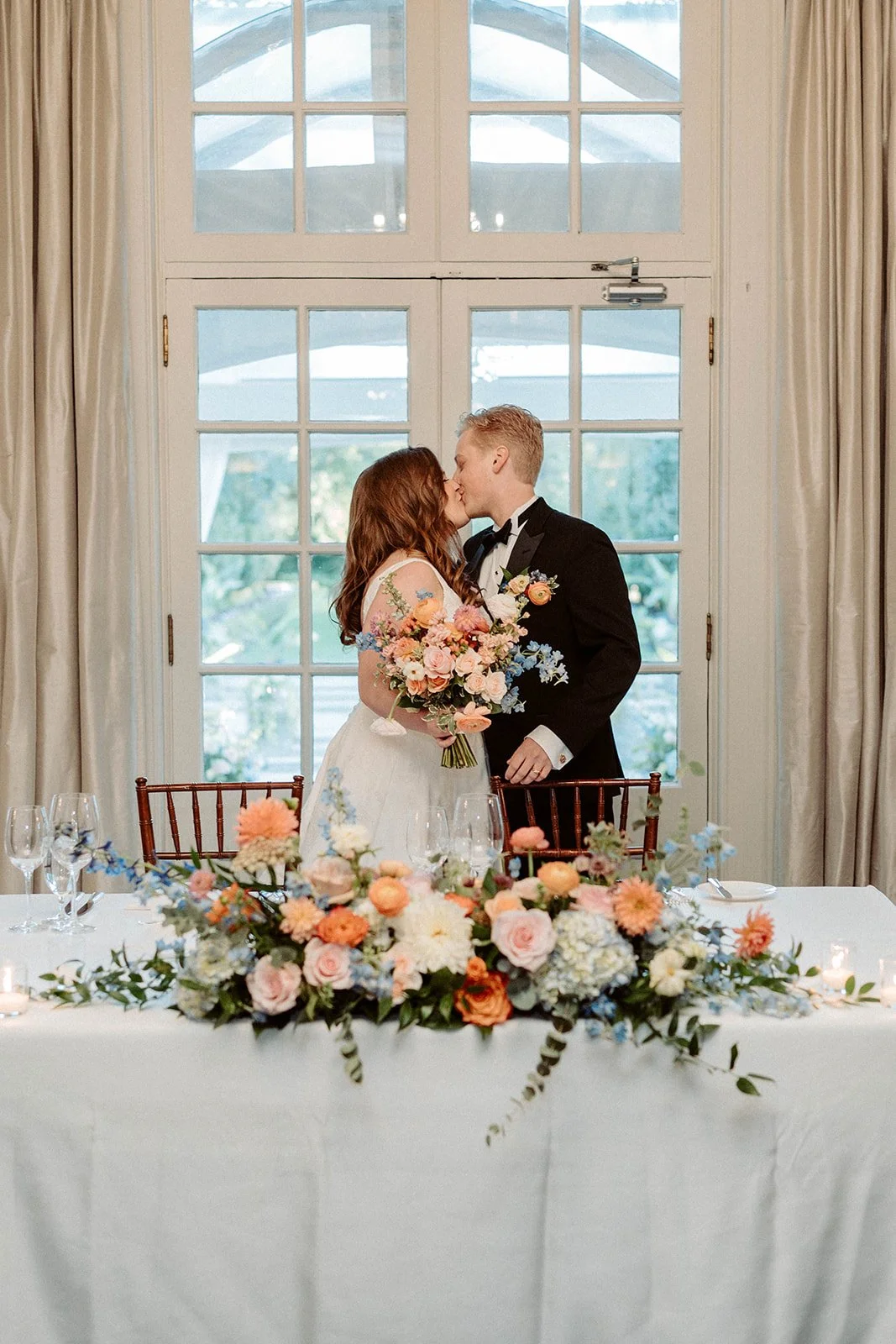 A bride and groom kiss behind a floral wedding table setup, with a large window and cream-colored curtains in the background.