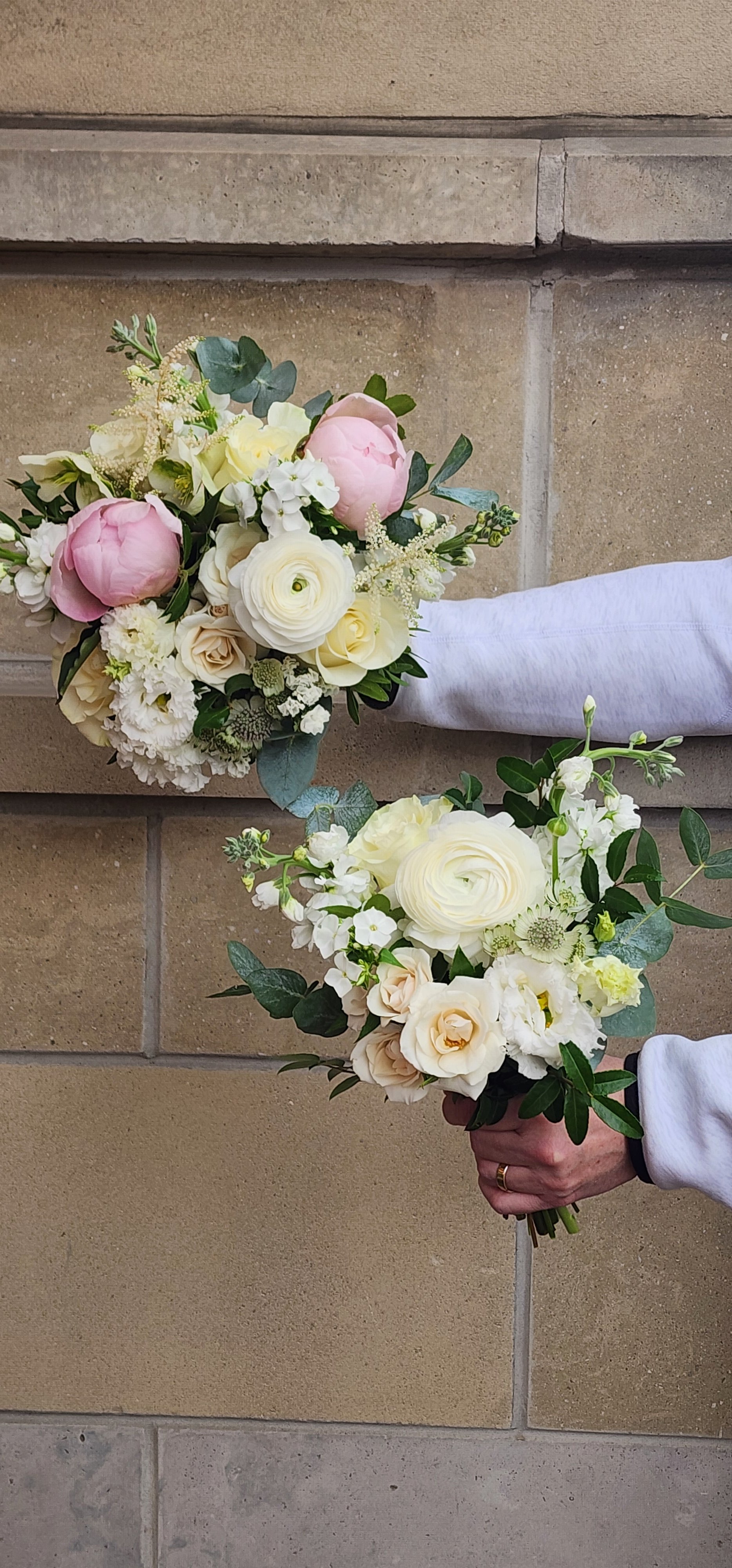 Two bouquets of white and pale pink flowers with green leaves, held by a person in a white sleeve against a tiled wall.