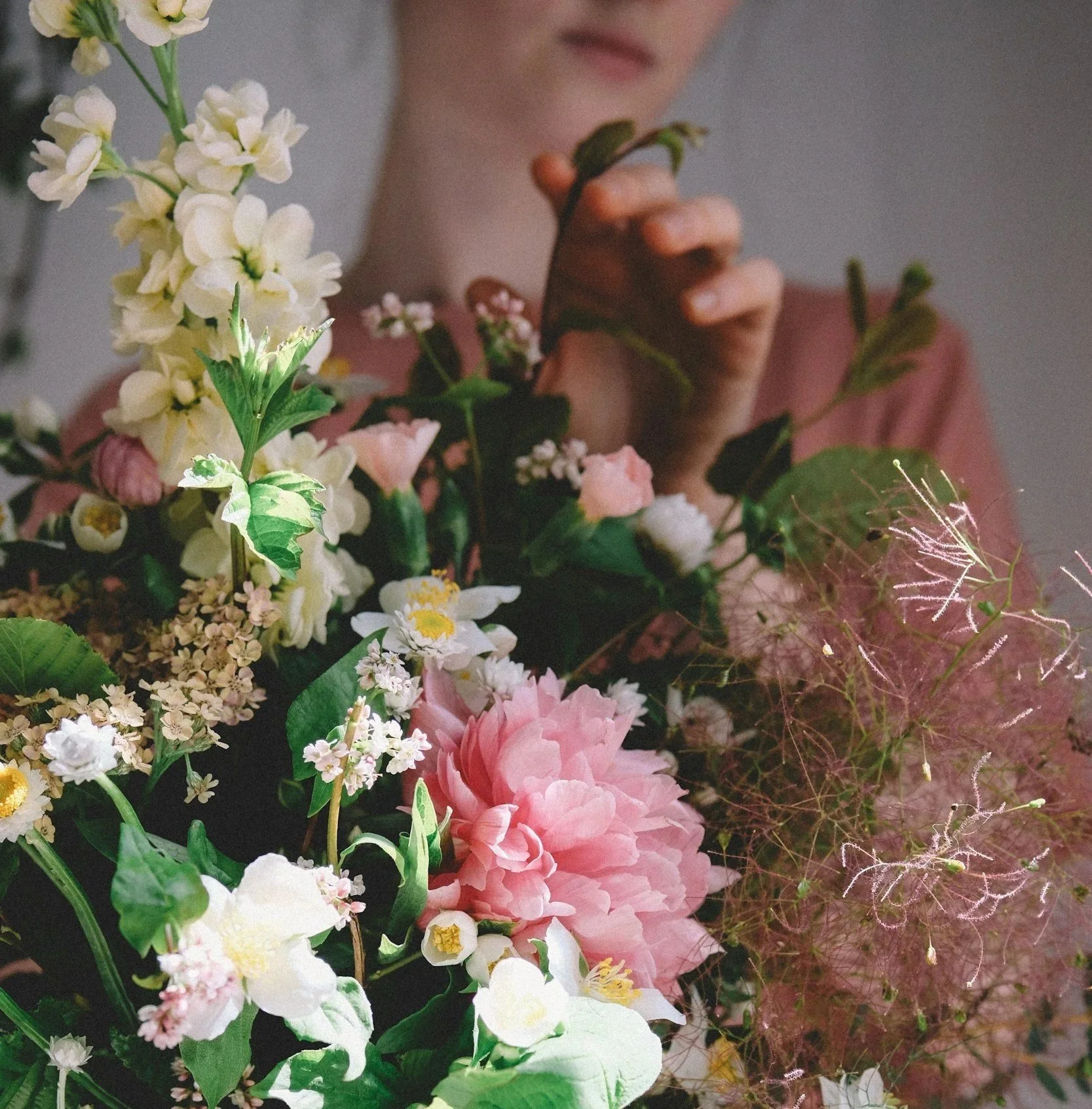 Close-up of a woman with short hair and a pink top arranging a colorful flower bouquet including pink peonies, white and yellow flowers, and green foliage.