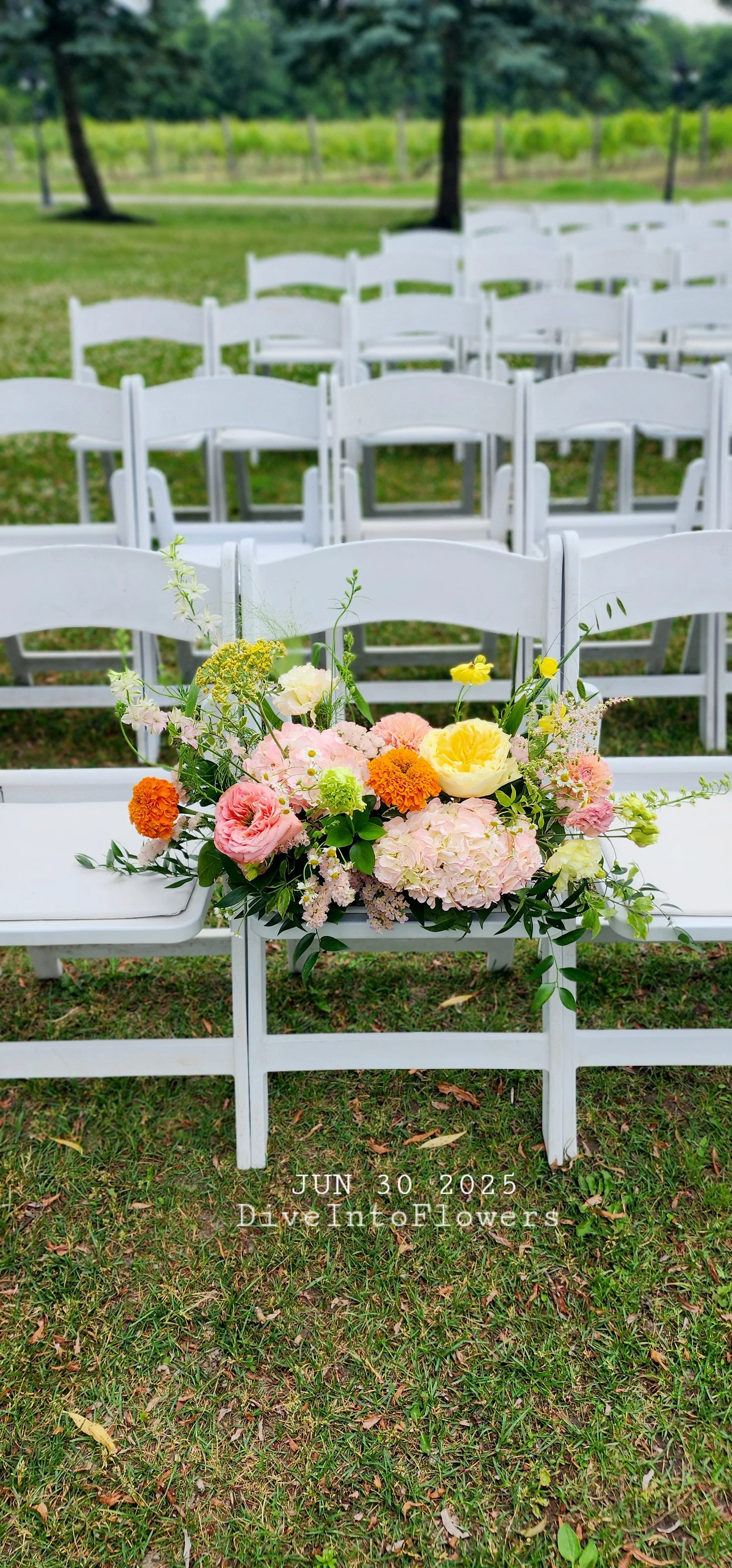 A white folding chair with a colorful flower arrangement on its seat, set on a grassy outdoor area with additional white chairs arranged in rows in the background, surrounded by trees. The photo has a date and website overlay at the bottom: June 30, 2025, DriveIntoFlowers.