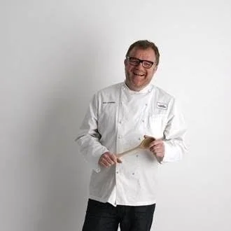 A smiling man in a white chef's coat holding a wooden spoon, standing against a plain white background.