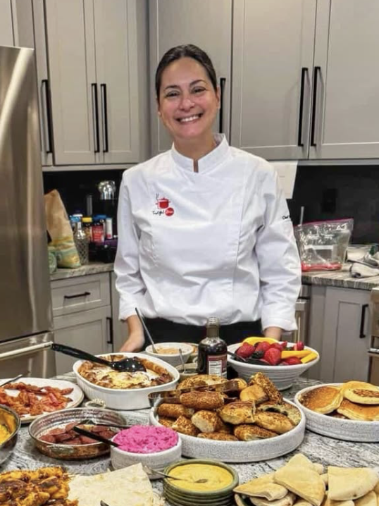 Smiling woman in a chef's coat standing behind a table filled with various dishes and desserts in a kitchen.