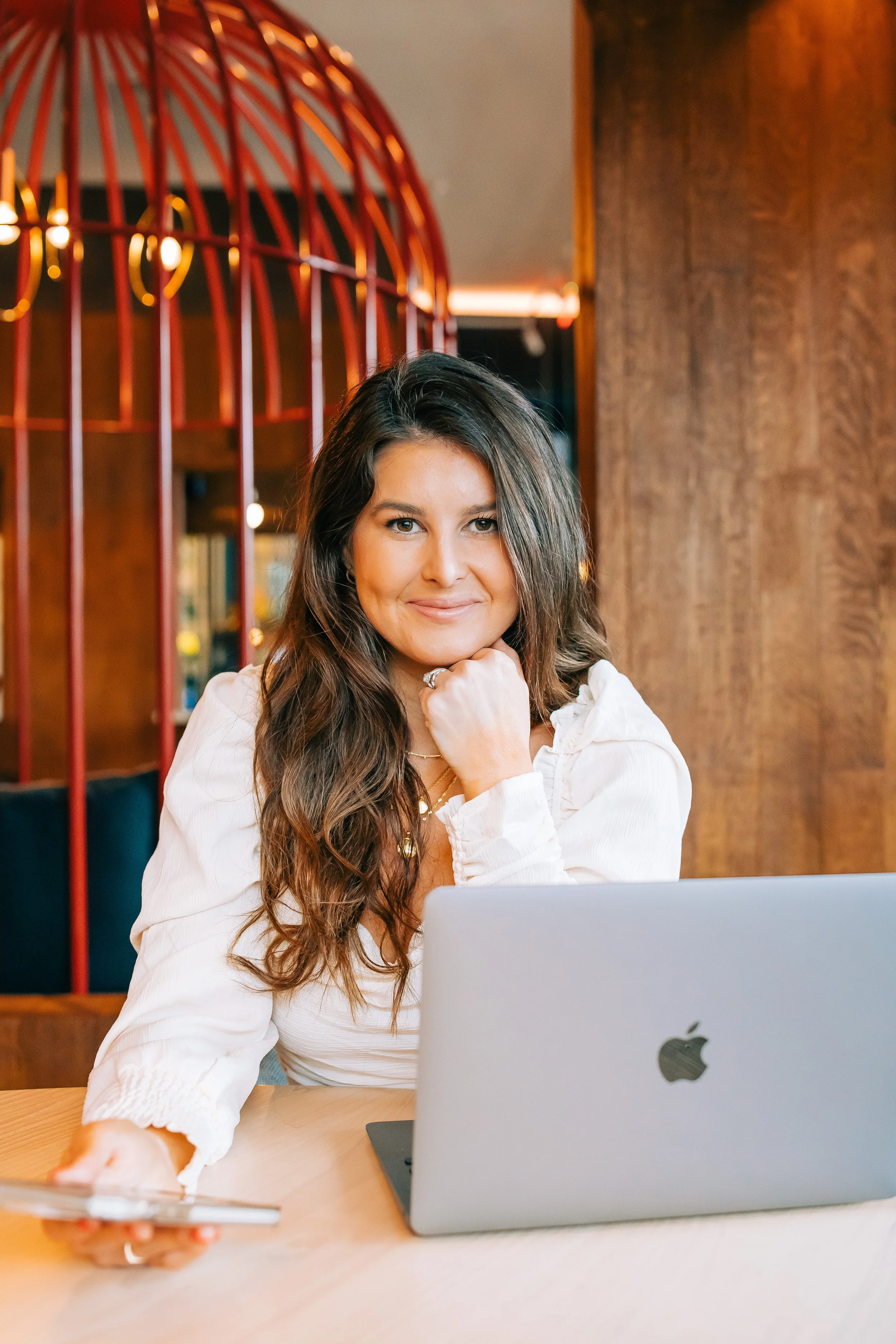 A woman with long wavy brown hair smiling at the camera, sitting at a table with an open silver Apple laptop, holding a smartphone in her right hand, inside a room with wooden walls and a red decorative arch.