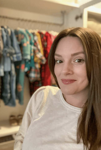 A woman with long brown hair taking a selfie in a room with a clothing rack in the background, featuring colorful jackets and sweaters.