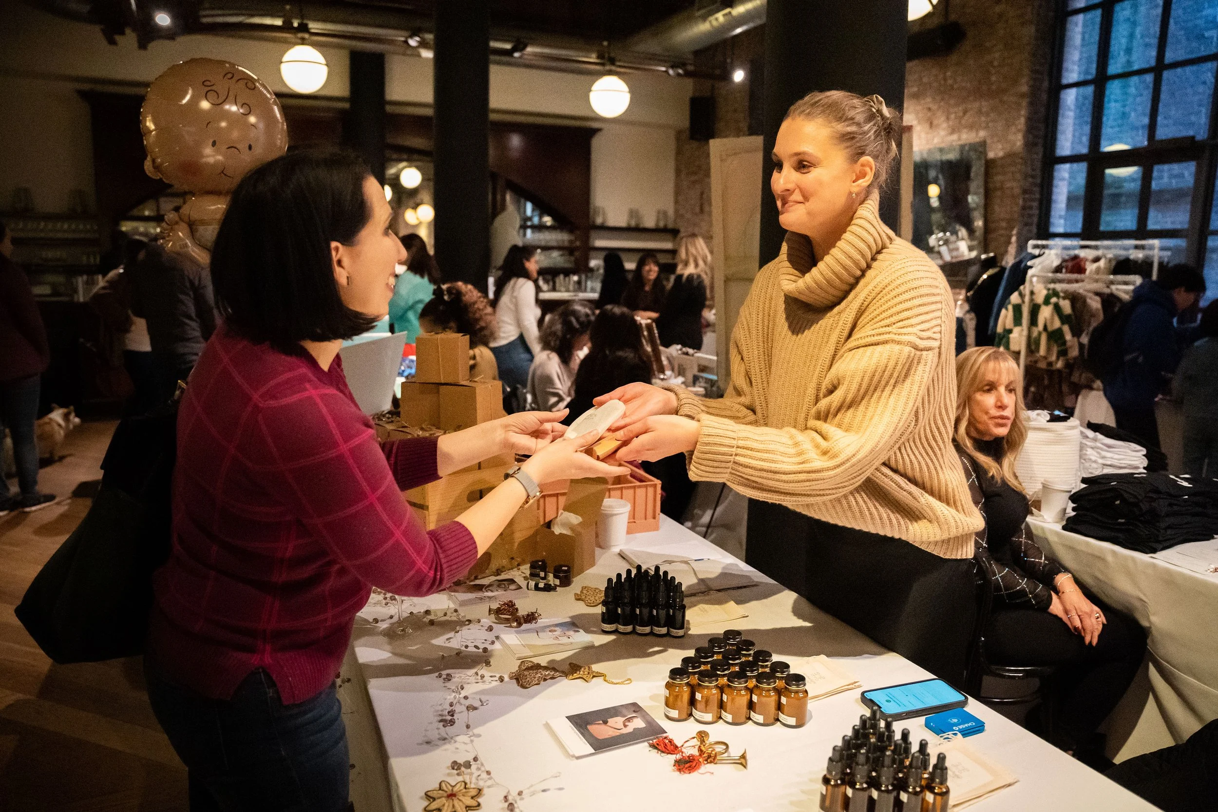 A woman in a beige sweater hands a small item to another woman in a red sweater at a market stall, with various small bottles and jewelry displayed on the table, in a busy indoor venue.