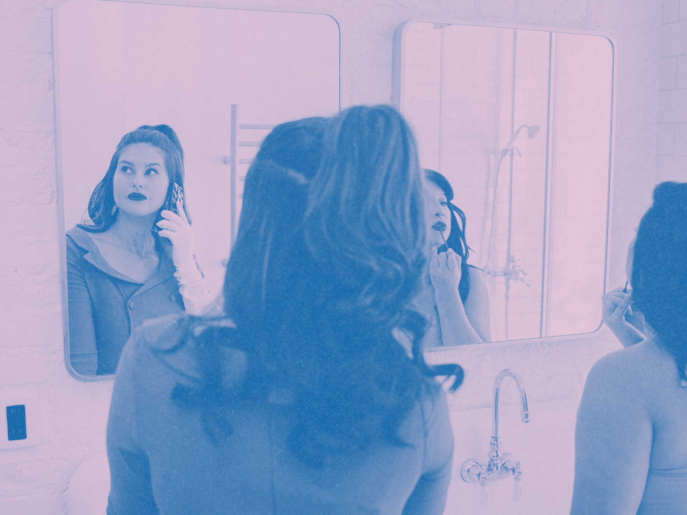 A woman in vintage clothing with dark lipstick and earrings looks into a mirror, talking on the phone. Two other women are applying makeup in front of a bathroom mirror, one of whom is applying lipstick.