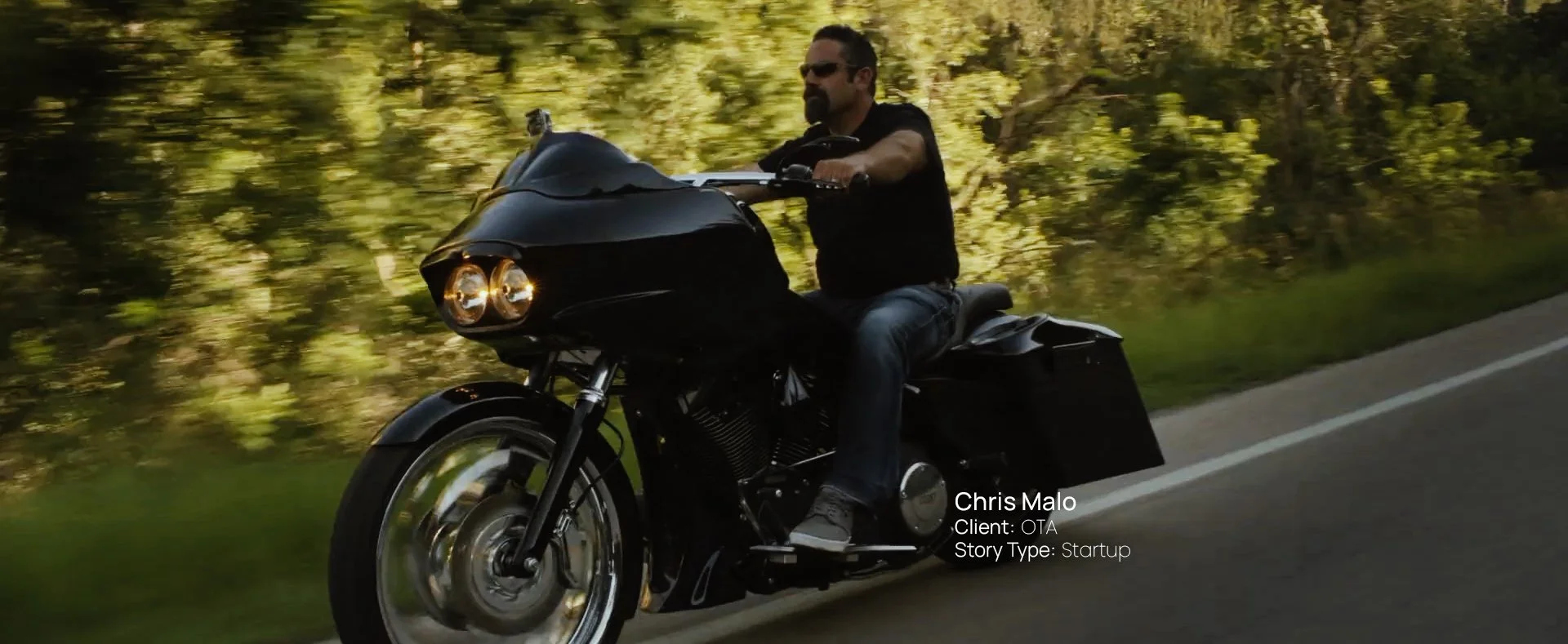 Man riding a black motorcycle down a tree-lined road in the Black Hills of South Dakota, still from a Passenger Productions startup documentary about Chris Malo