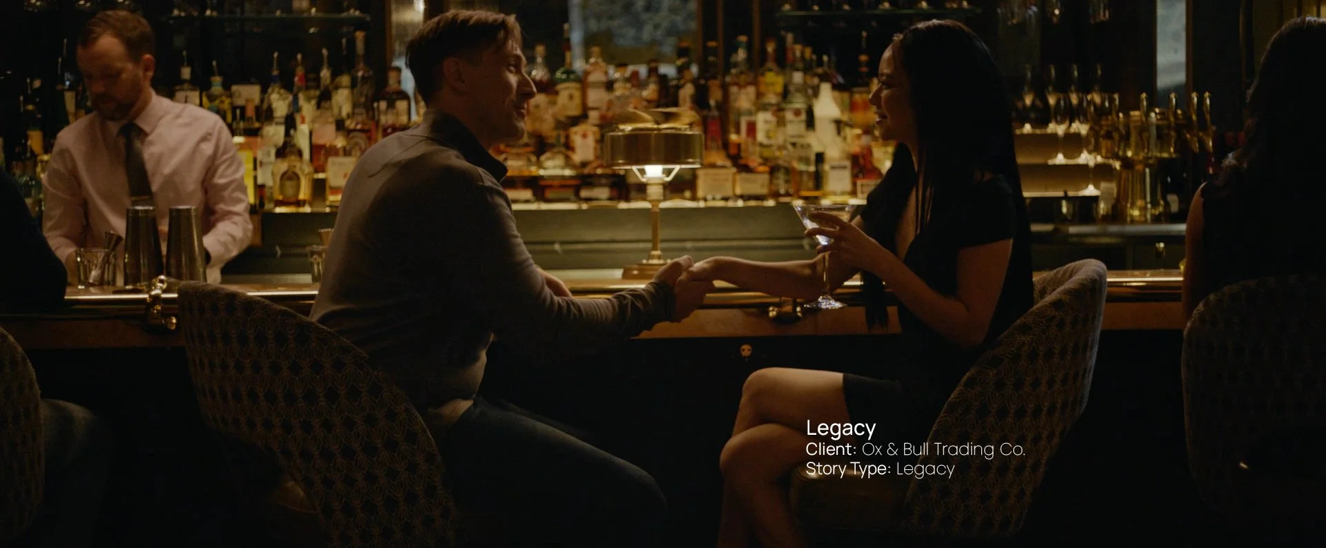 Man seated at a bar meets woman and shakes hand surrounded by bottles, still from a Passenger Productions legacy documentary for Ox & Bull Trading Co.