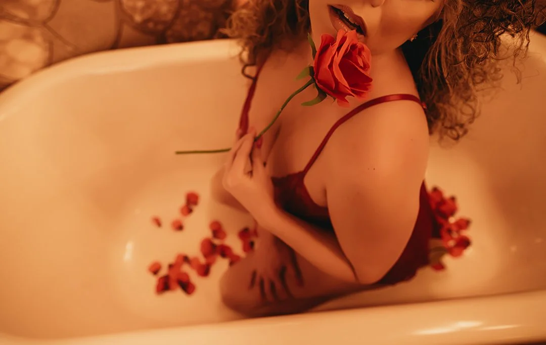 A woman with curly hair in a red dress inside a bathtub, holding a red rose in her mouth, surrounded by rose petals.