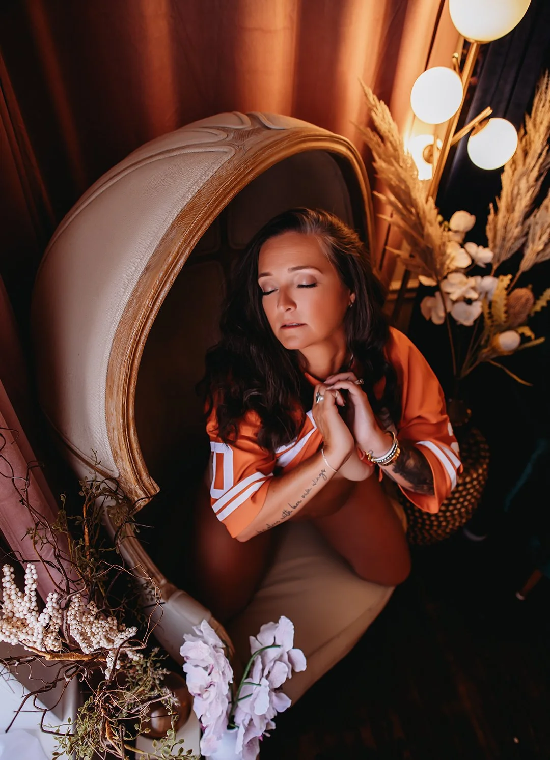 A woman with long dark hair and tattoos on her arms, sitting on a beige velvet chair with a rounded back, eyes closed, and hands clasped near her chin. She wears an orange sports jersey and jewelry. The setting is warm with soft lighting, beige curtains, dried pampas grass, and flowers.