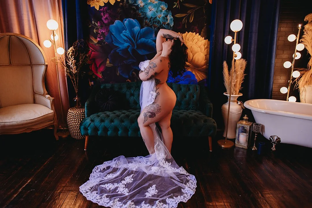 A woman with tattoos standing seductively in front of a dark green tufted sofa, holding a sheer lingerie dress, surrounded by stylish decor including a floral wall, vases, lamps, and a vintage bathtub.