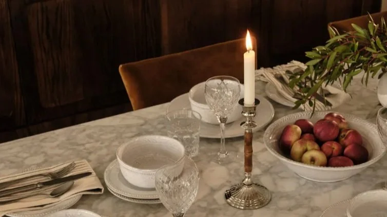 A dining table set with white marble top, featuring plates, bowls, crystal glasses, a lit candle in a candlestick holder, a bowl of apples, and a vase with green leaves.