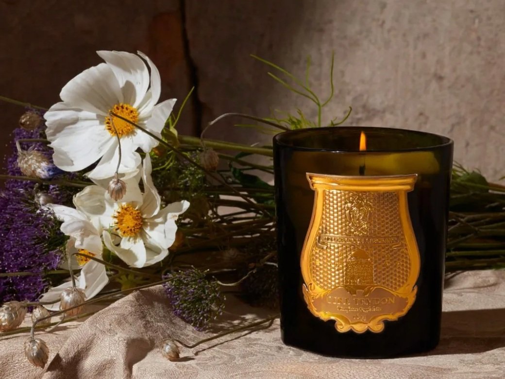 A lit candle in a black glass holder with a golden crest emblem, lying on a beige cloth, with white flowers and purple and green plants in the background.