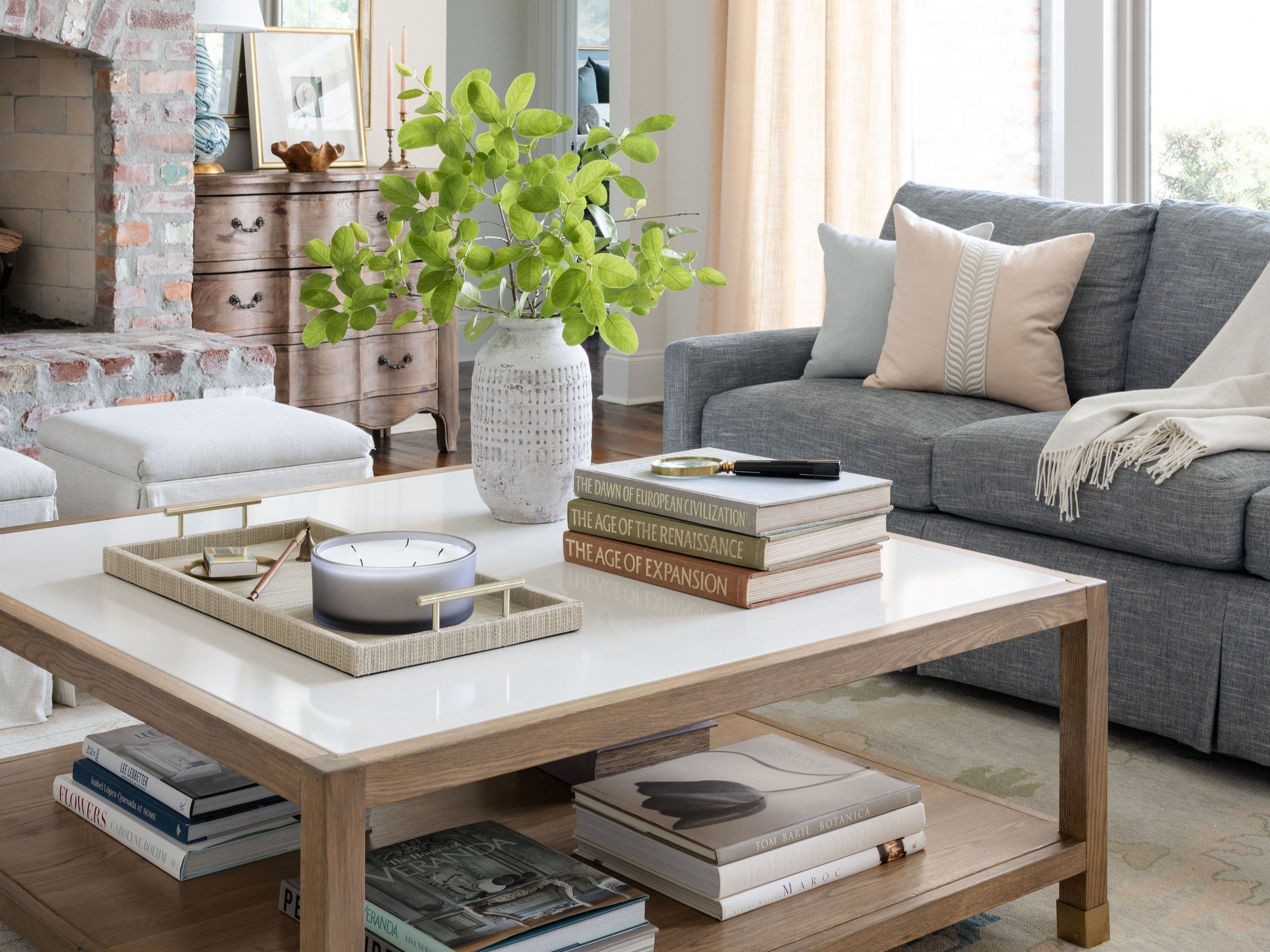 A cozy living room with a gray sofa, beige and cream pillows, a wooden coffee table with books and a tray, a large white vase with green leafy plant, a brick fireplace, and light streaming through large windows with beige curtains.