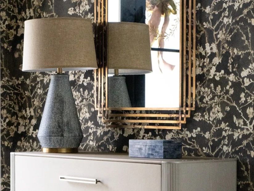 Interior view featuring a table lamp with a beige lampshade, a decorative wall mirror with a gold geometric frame, a patterned black and gold wallpaper, and a white cabinet with a gray stone box on top.