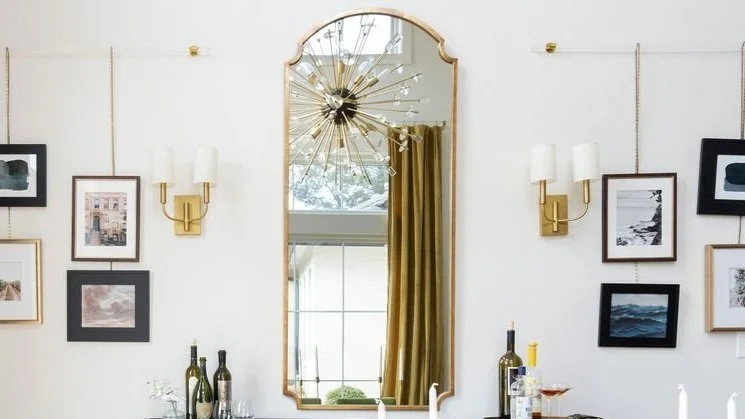 A wall with a large arched mirror in the center, surrounded by framed artwork and two wall-mounted gold and white lamps. A chandelier with multiple gold spokes and bulbs hangs above, and a window with a yellow curtain is reflected in the mirror.