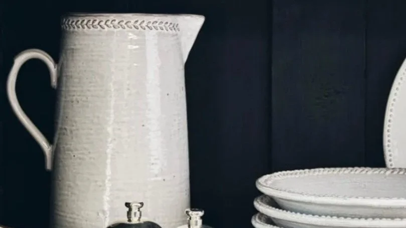 A white ceramic pitcher, white plates stacked, dark background.