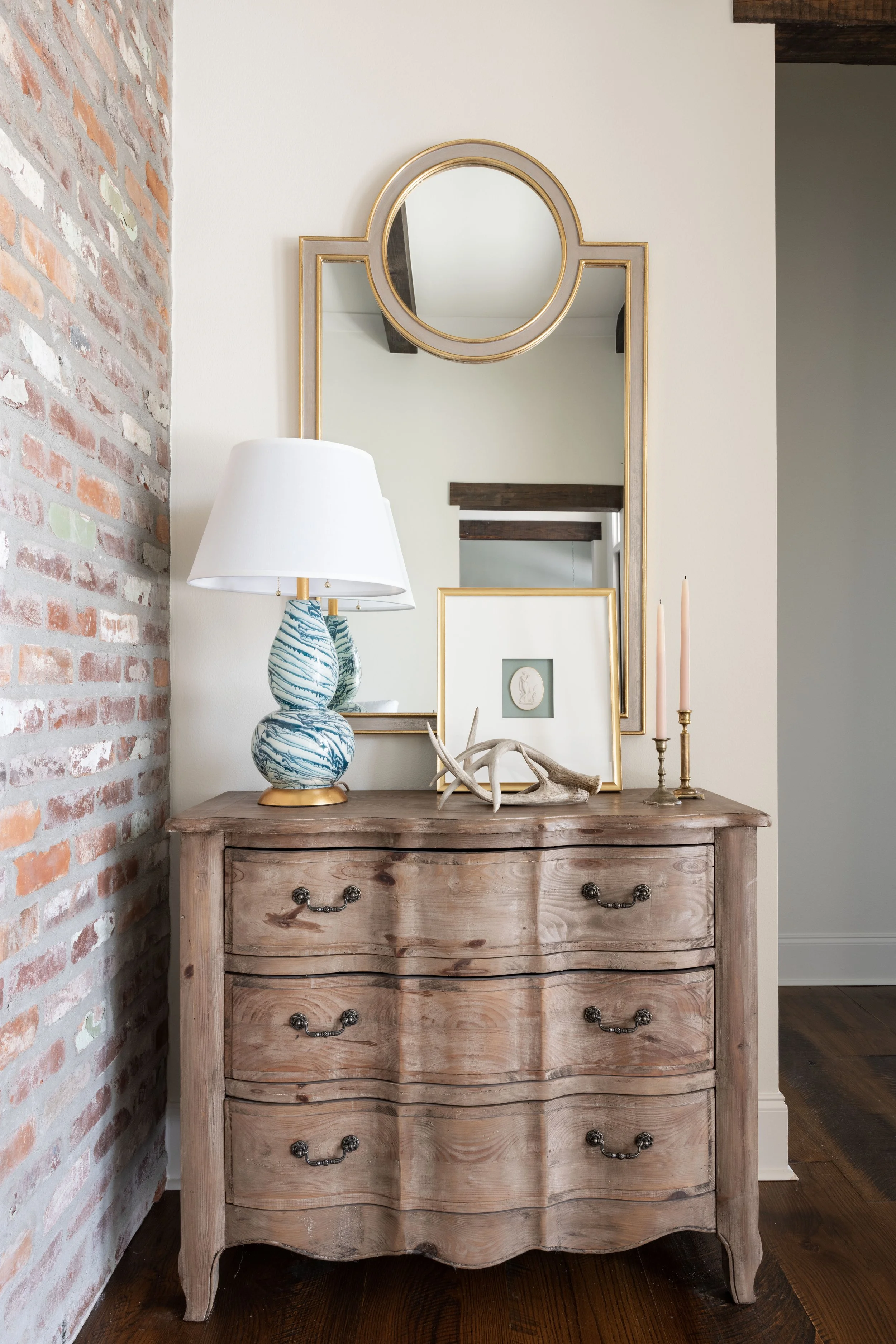 A rustic wooden dresser with ornate handles, topped with a white lampshade lamp, a framed picture, antlers, and two candlesticks. A large mirror with a gold frame hangs above it, reflecting the room's ceiling beams and part of a doorway.