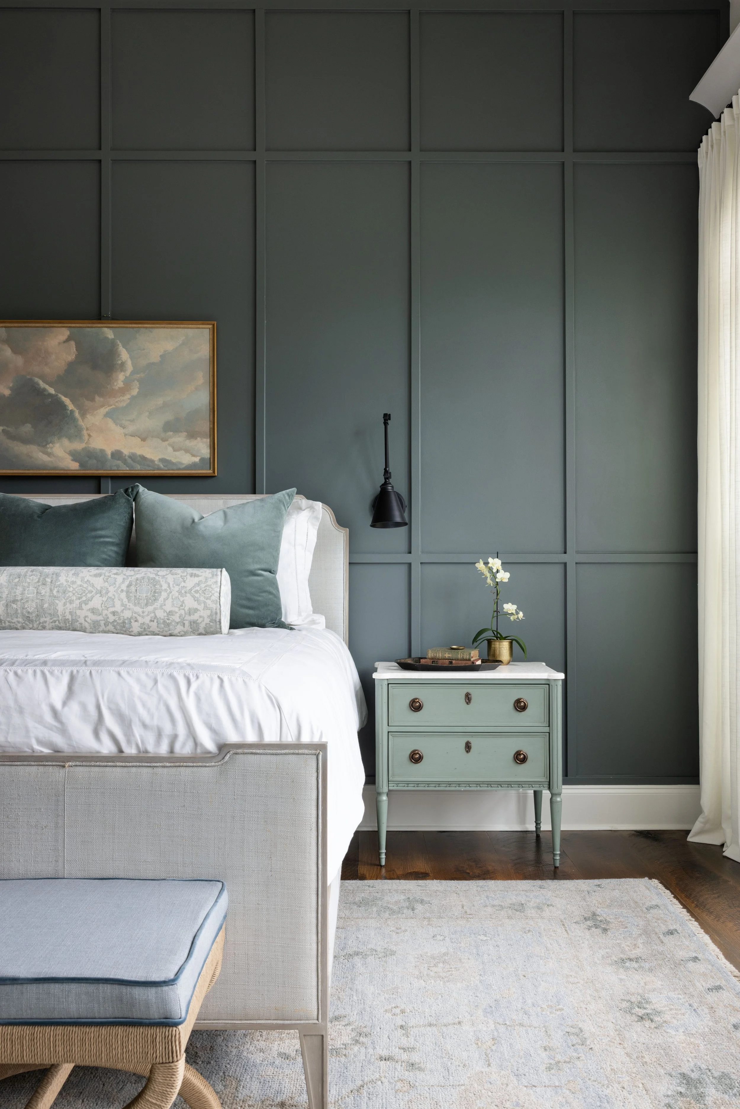 A bedroom corner with a bed, pillows, a nightstand with a flower, and a dark green paneled wall.