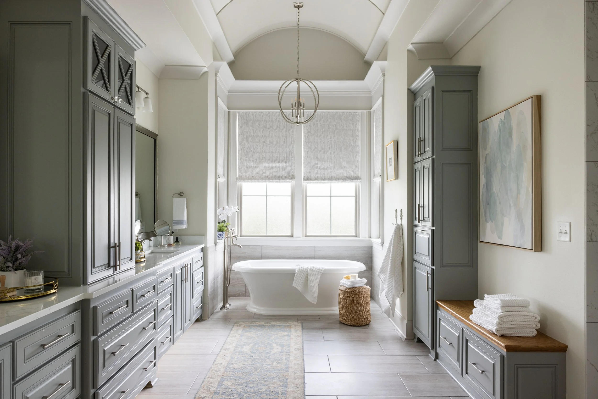 Bright and spacious bathroom with gray cabinetry, a freestanding bathtub near large windows, and a neutral color palette with decorative accessories and artwork.