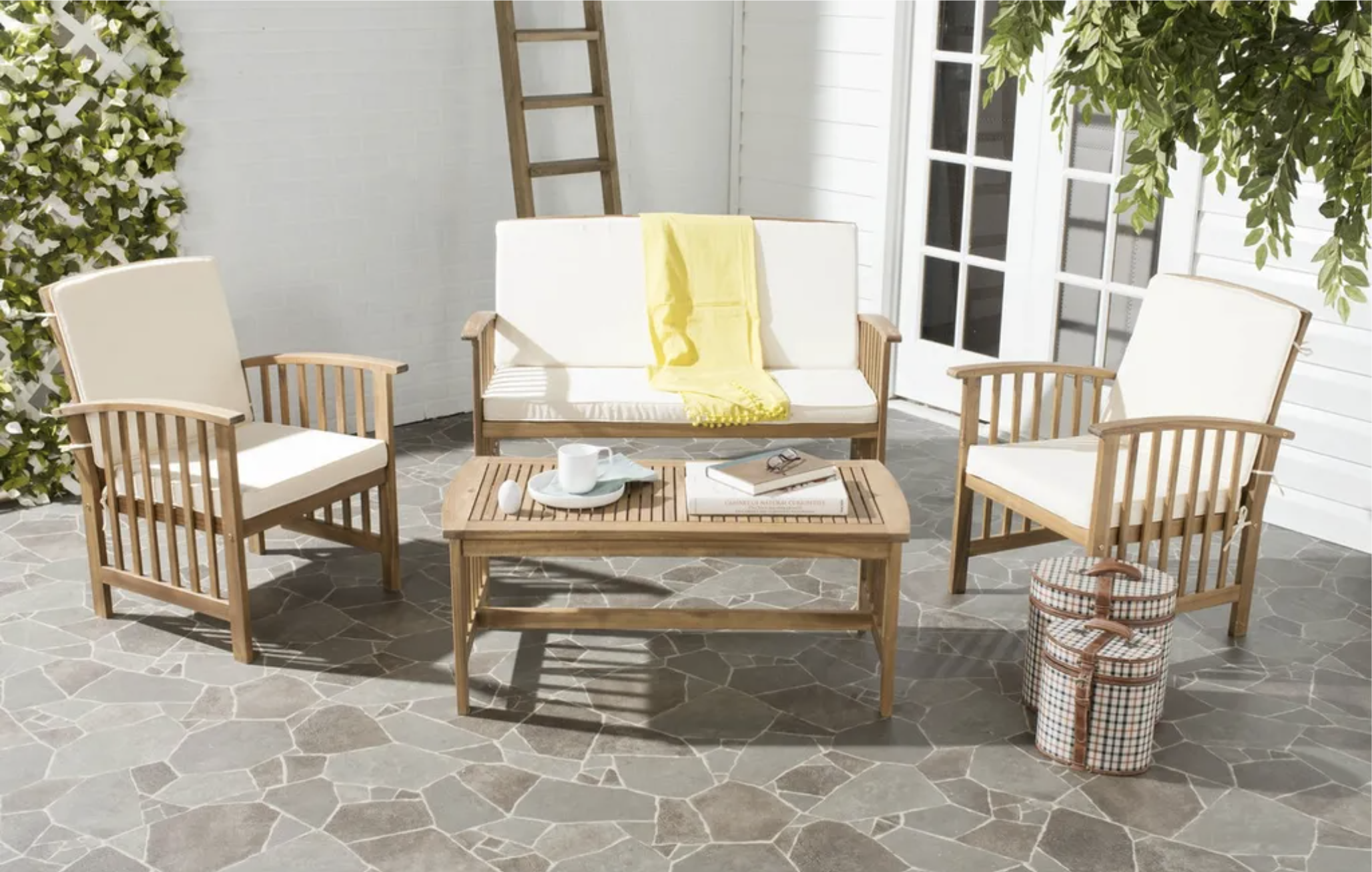 Outdoor patio with white cushioned wooden furniture, a coffee table with a cup and books, and decorative luggage, surrounded by greenery and a white brick wall.