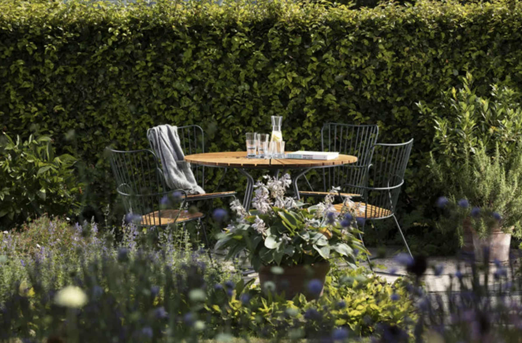 A round wooden outdoor table with four black metal chairs, one with a gray blanket, arranged in a garden with lush green shrubs and plants; items on the table include a glass pitcher, two glasses, and a book.