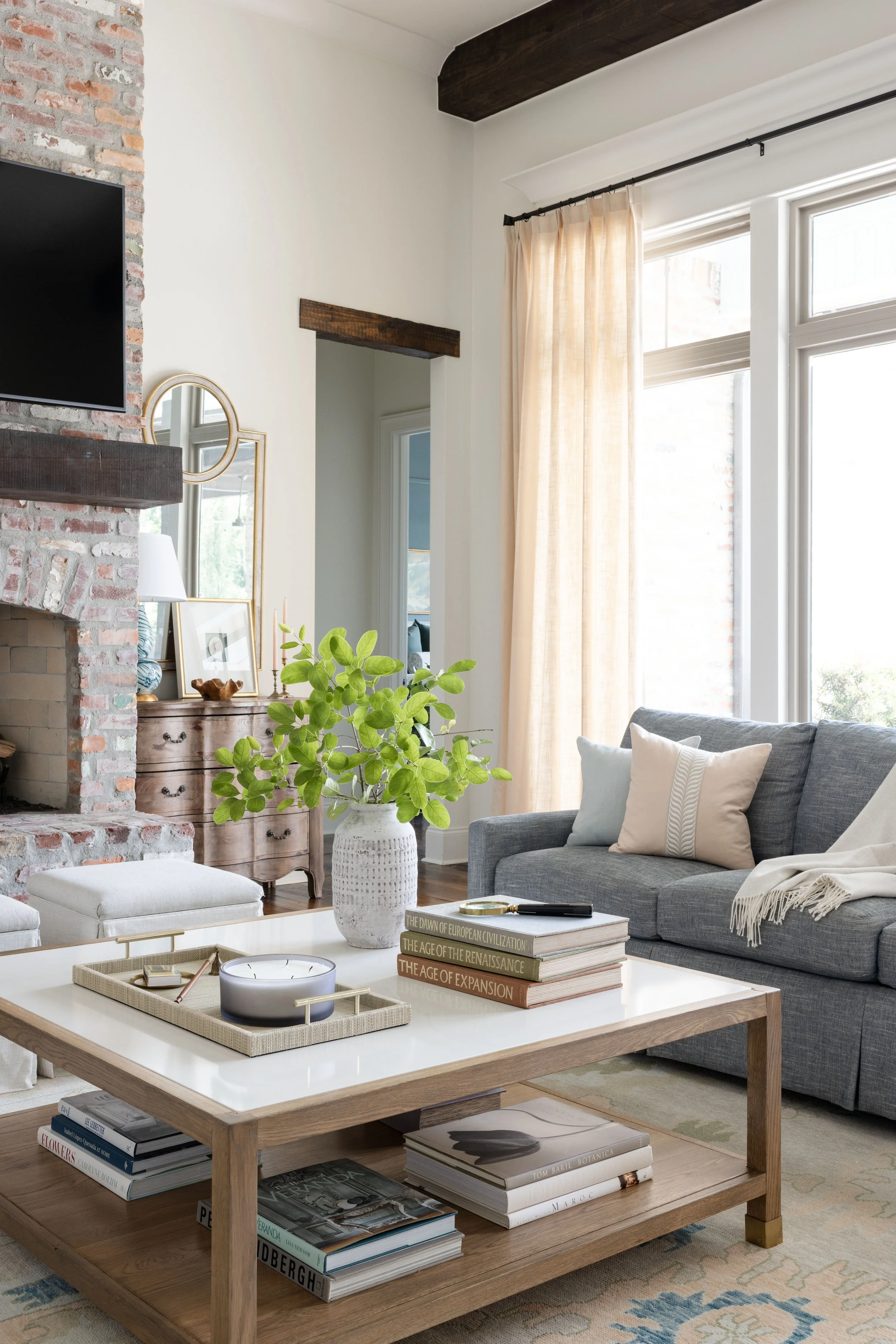 A cozy living room with a stone fireplace, a wall-mounted TV, a wooden coffee table with books and decorative items, a gray couch with cushions and a throw blanket, and a large window with sheer curtains.