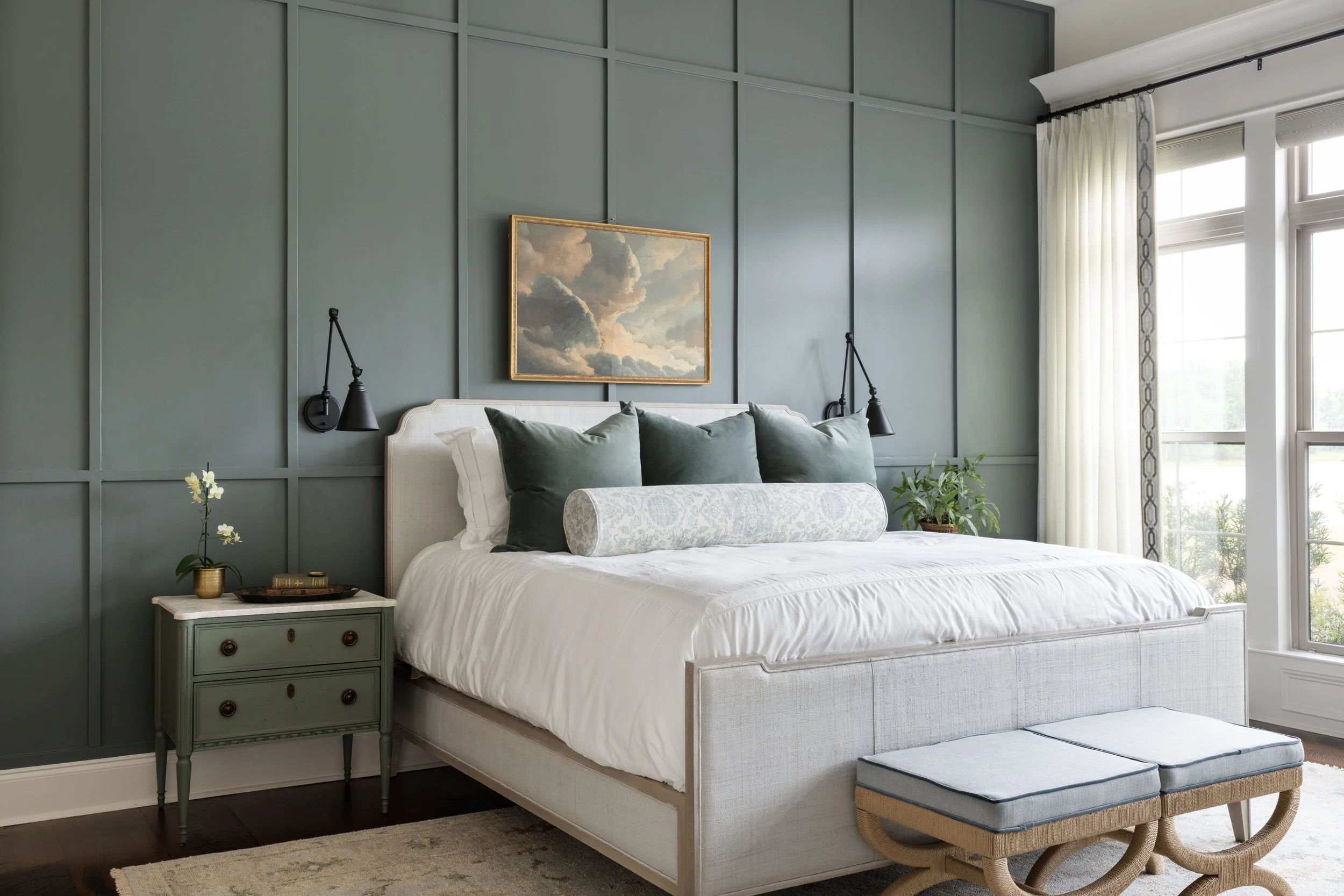 A bedroom with a green paneled wall, a white bed with green and white pillows, a small nightstand with a plant, and a bench at the foot of the bed. There is a window with white curtains on the right.