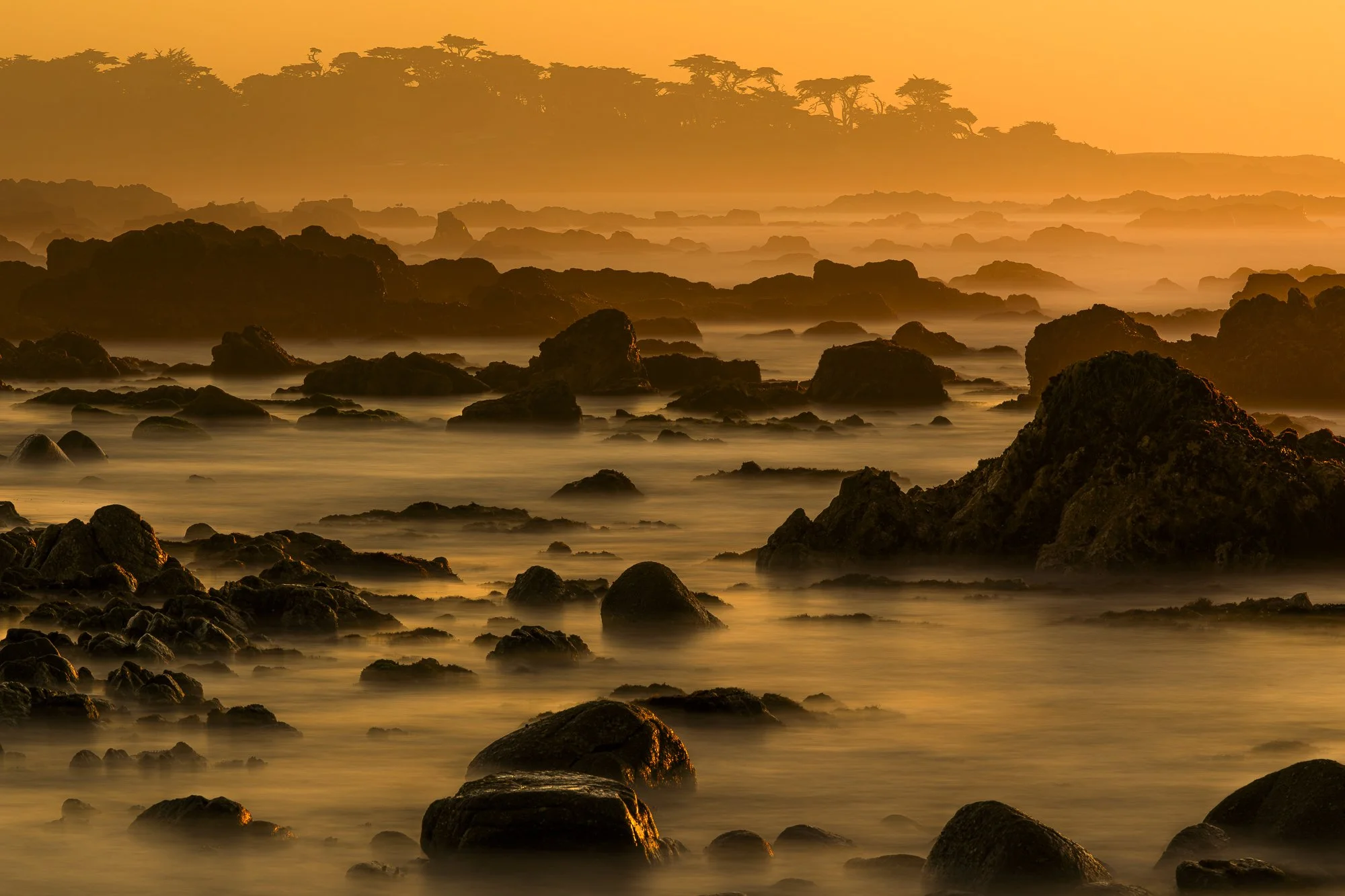 Golden sunset over the rocky shoreline of Pebble Beach with misty waves and coastal haze.
