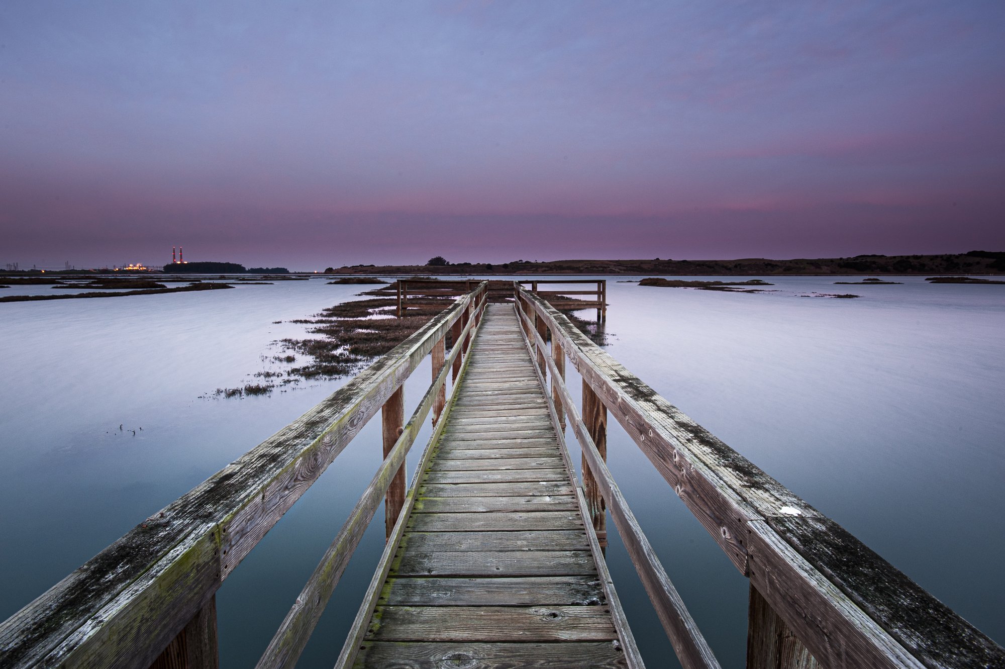 Wooden footbridge over still waters at Elkhorn Slough at dusk, with purple sky, by Jason Bradley.