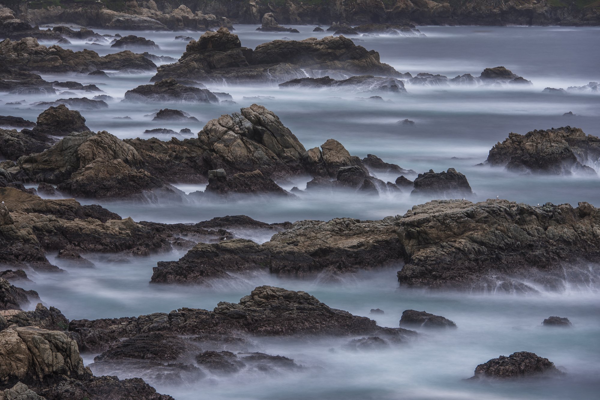 Long exposure of waves flowing around rugged coastal rocks under fog along the Pacific coast by Jason Bradley.
