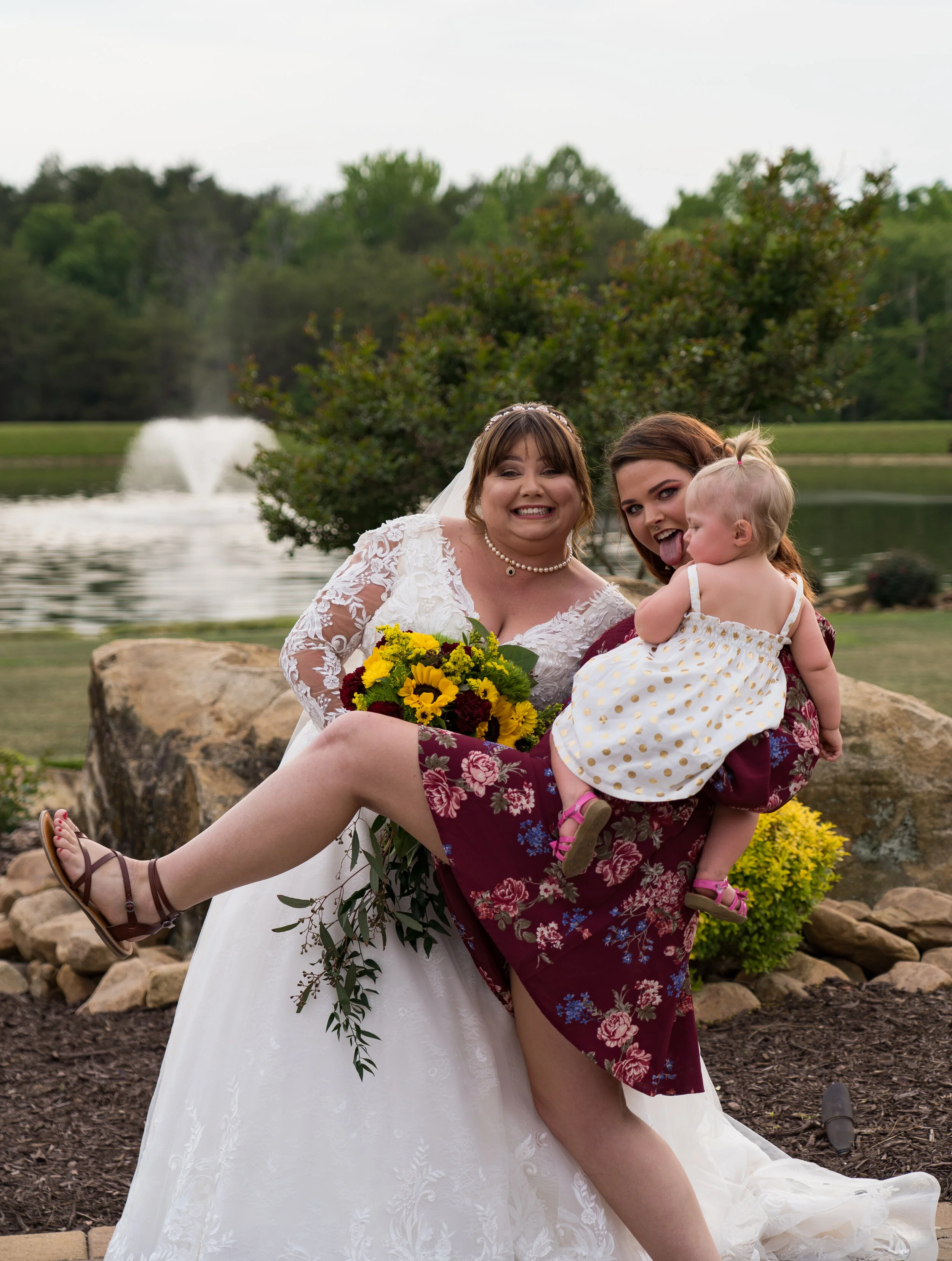 Three women, including a bride in a wedding dress, all smiling near a pond with a fountain and trees in the background. One woman is holding a little girl in a white dress with gold dots.