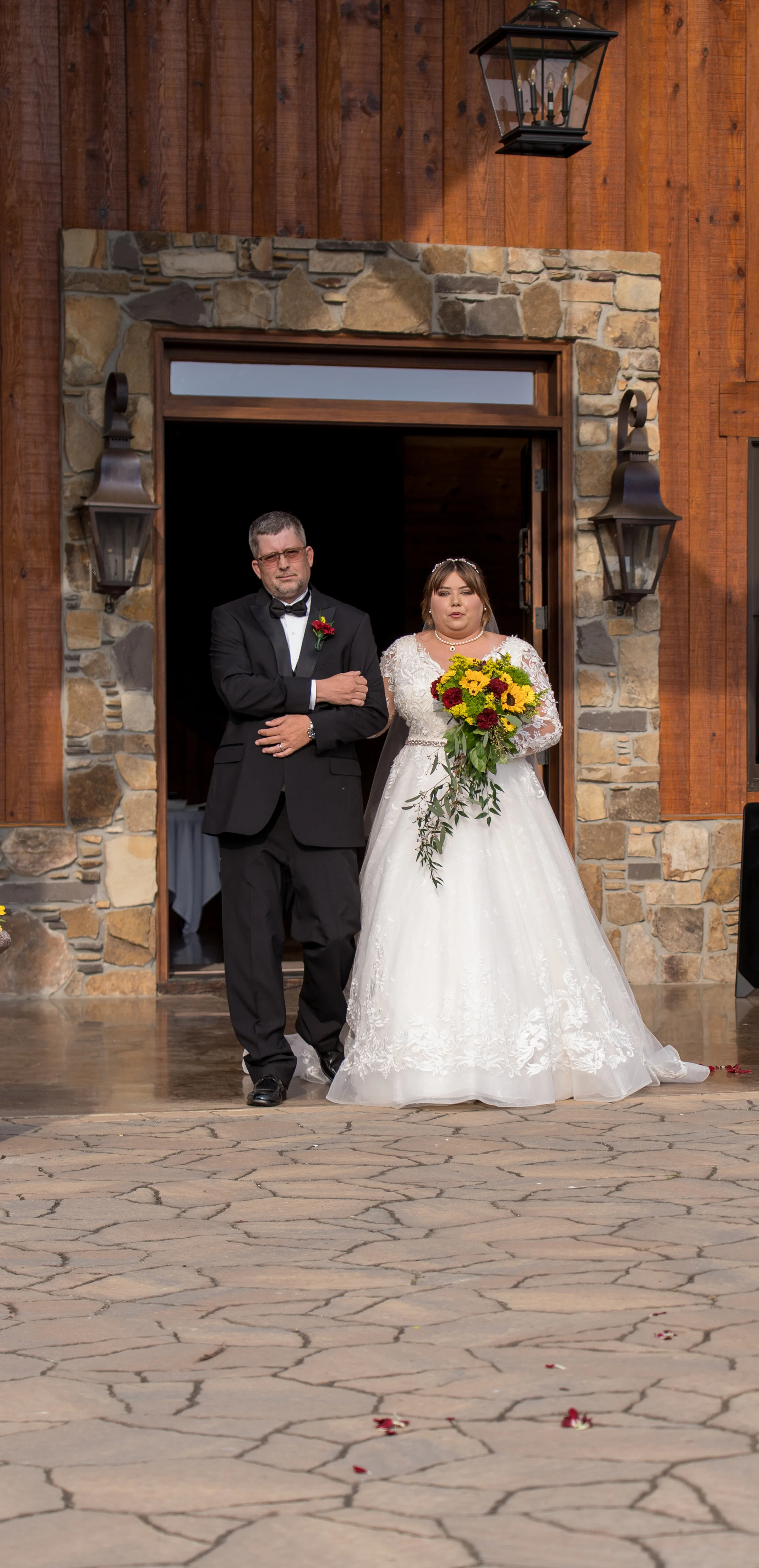 A bride holding a bouquet of yellow, red, and green flowers, standing next to a man dressed in a black tuxedo, inside the doorway of a rustic building with stone and wood exterior, during a wedding ceremony.