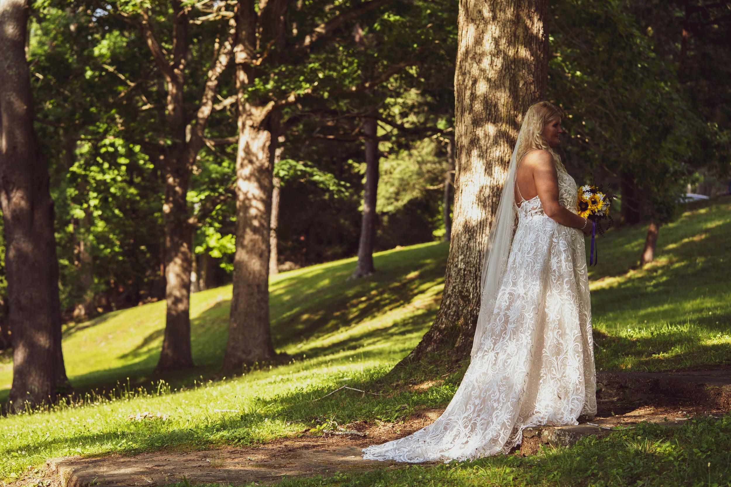 Bride in a white lace wedding dress with a long train standing outdoors in a wooded area, holding a bouquet of sunflowers, and posing near a tree.