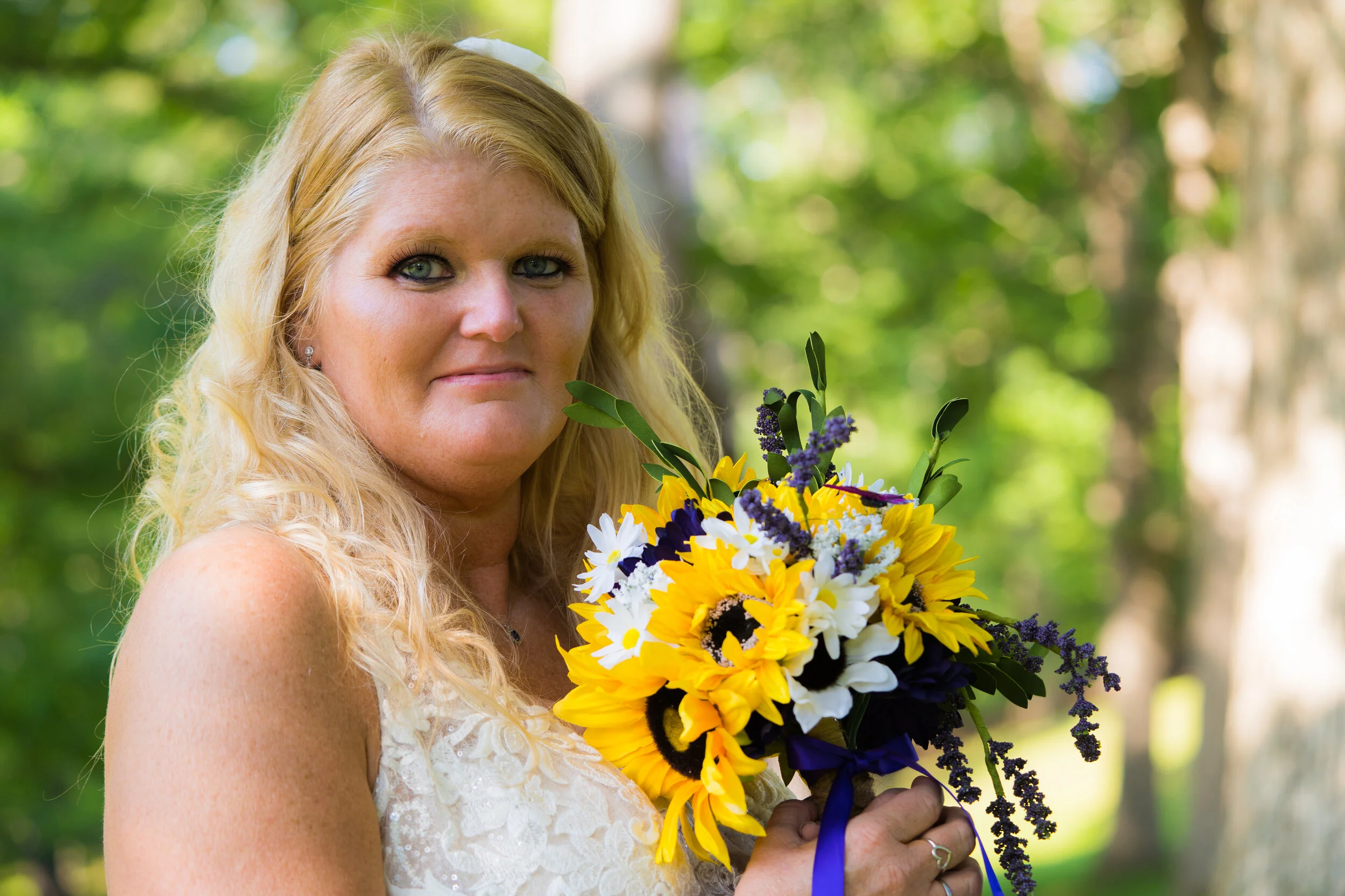 A blonde woman in a white lace dress holding a bouquet of yellow, white, and purple flowers outdoors in a green, wooded area.