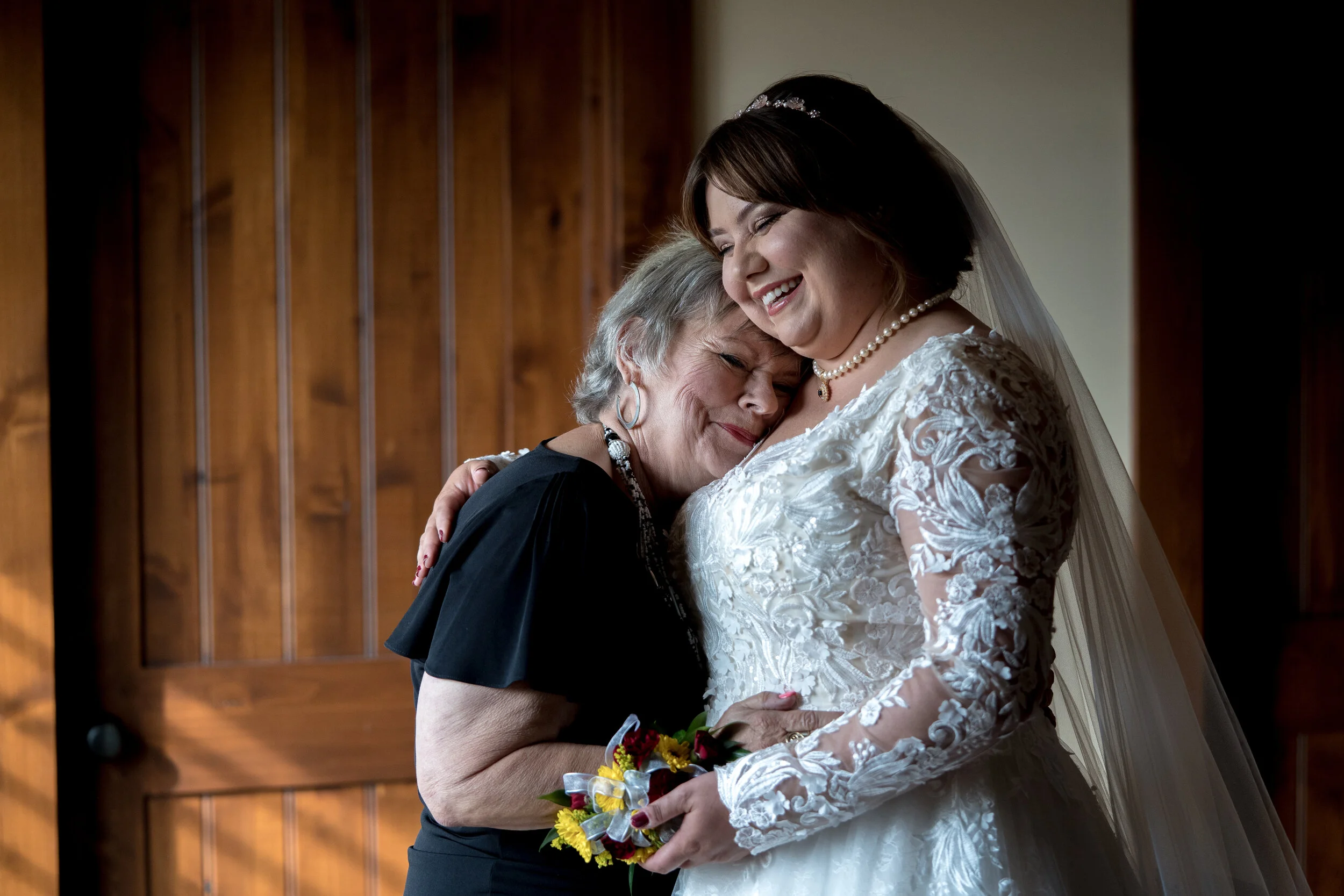 A bride in a white lace wedding dress, holding a bouquet, hugging an elderly woman in a black dress, both smiling affectionately, inside a room with wooden walls.
