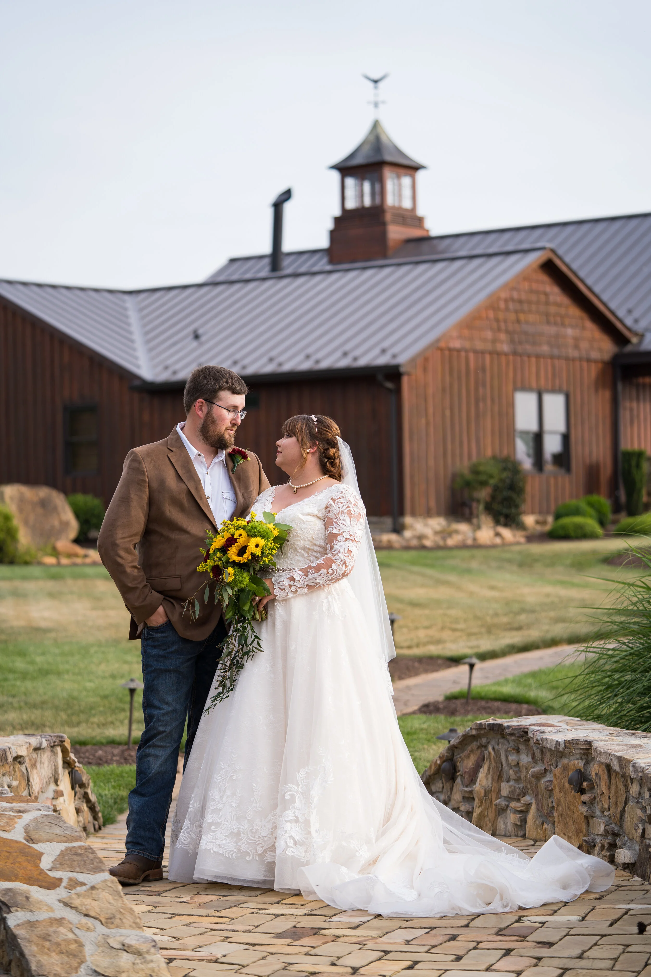 A bride and groom standing together outdoors on a wedding day, gazing into each other's eyes. The bride is holding a bouquet of sunflowers, and they are standing on a stone pathway with a rustic wooden building in the background.