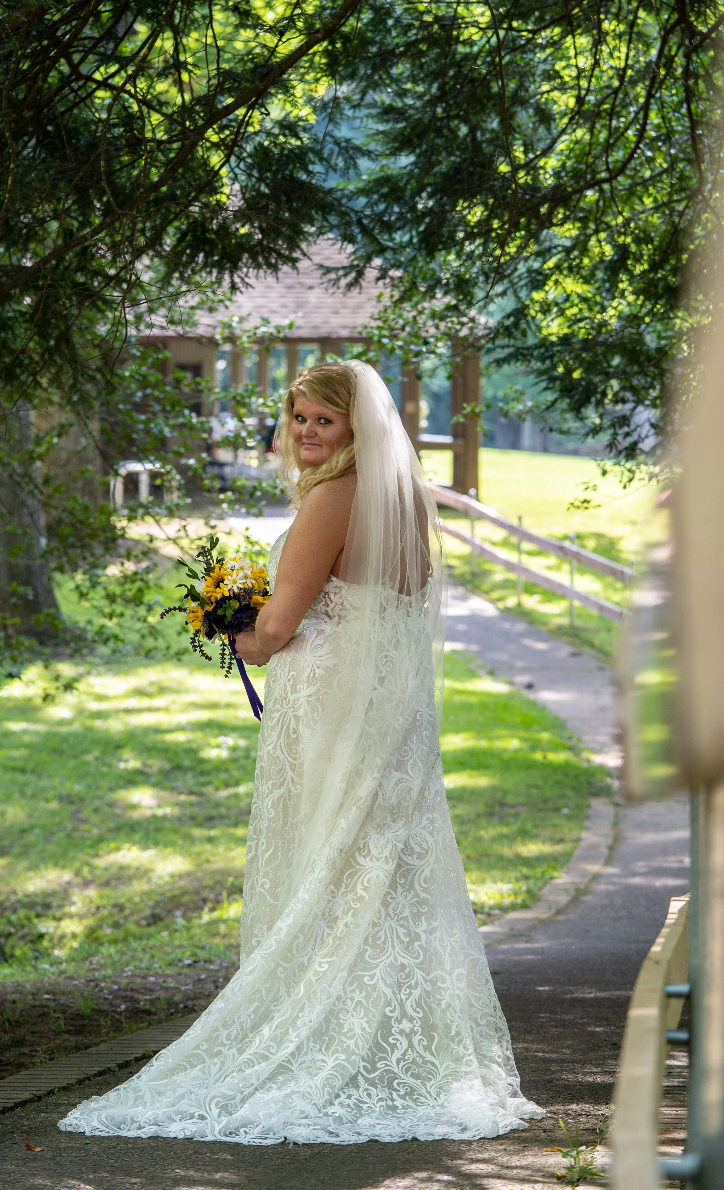 A bride in a white lace wedding dress holding a bouquet of yellow and white flowers standing outdoors under trees with dappled sunlight.