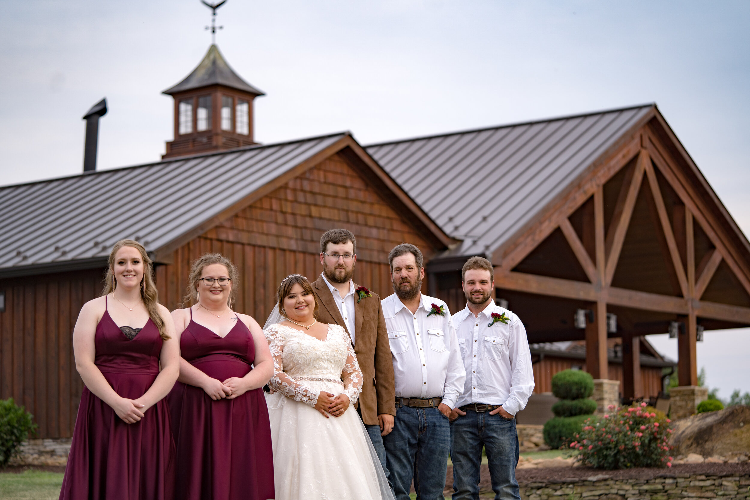 Group of six people standing in front of a wooden building, possibly a barn, during a wedding. The bride is in the center wearing a white lace wedding dress, and the groom is beside her in a brown jacket. Two women in maroon dresses and two men in wh