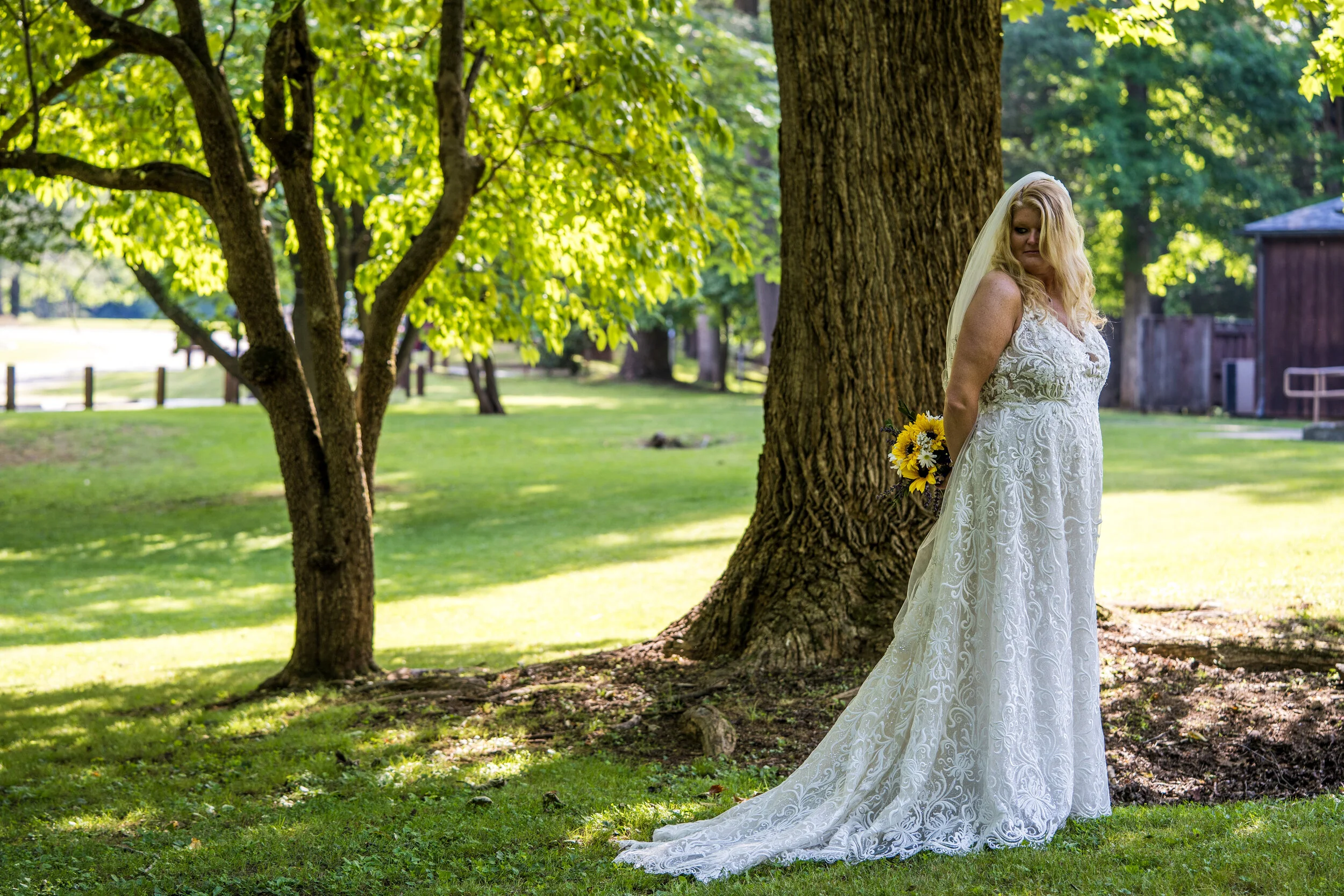 A woman in a white lace wedding dress holding a bouquet of sunflowers and wildflowers, standing outdoors near a large tree, with green grass and trees in the background.