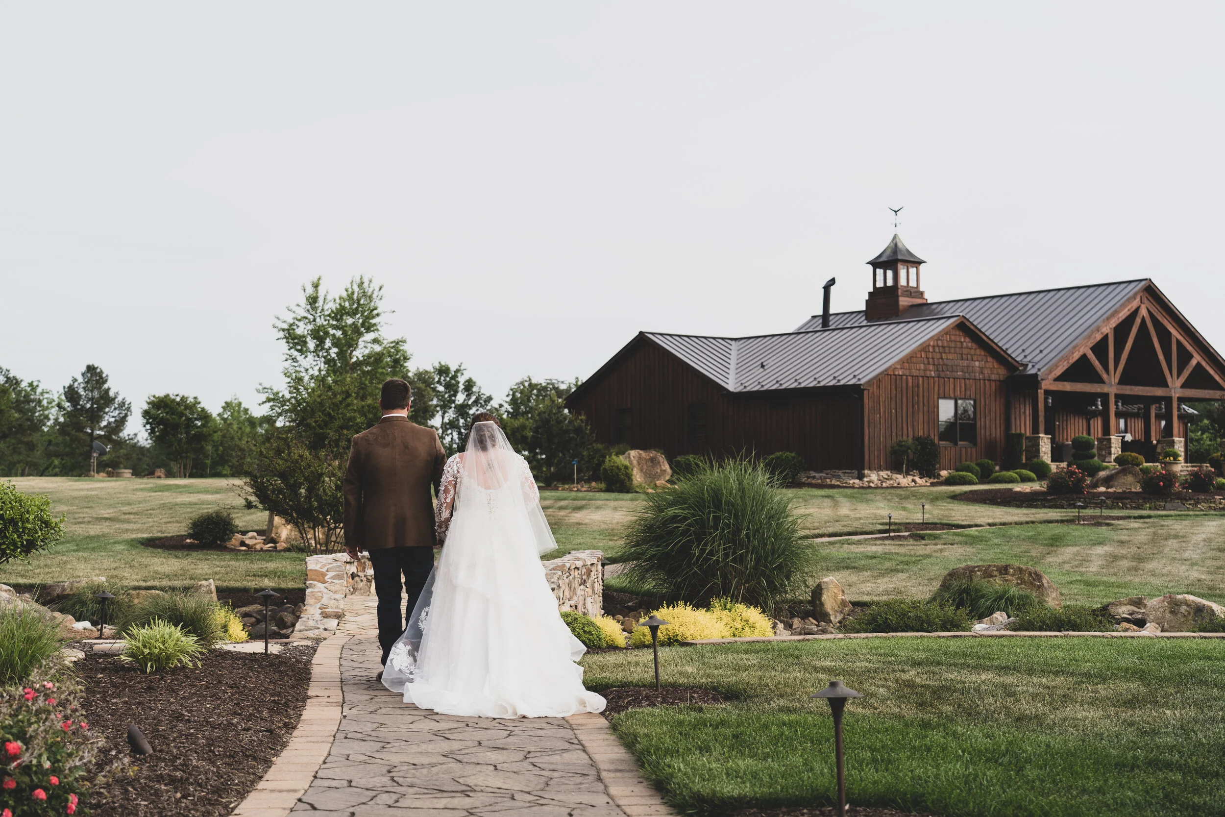 Bride and groom walking on a stone pathway towards a rustic barn in a landscaped garden.