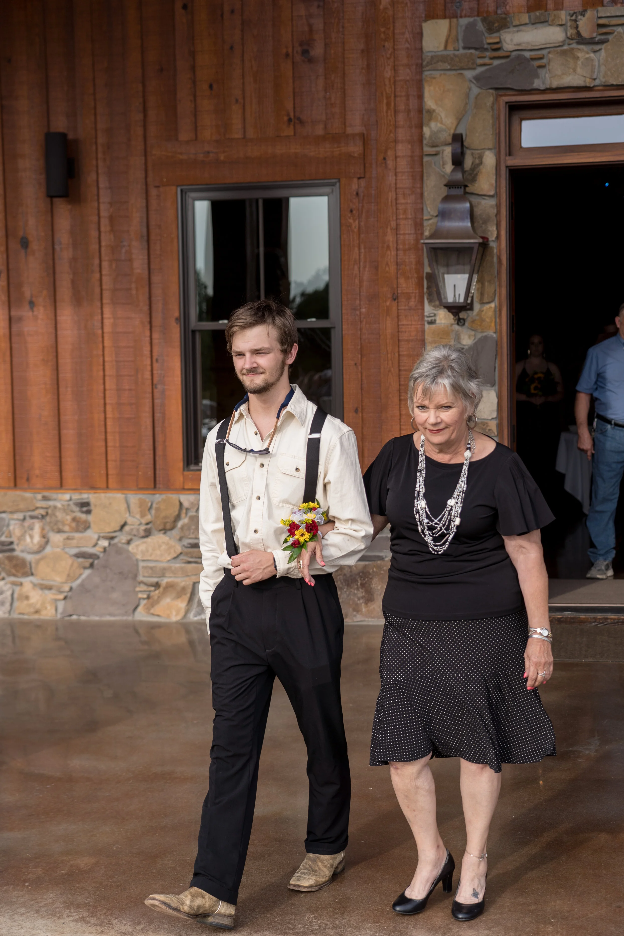 A young man with suspenders and a girl with a corsage walking together during a wedding event outside a rustic venue.