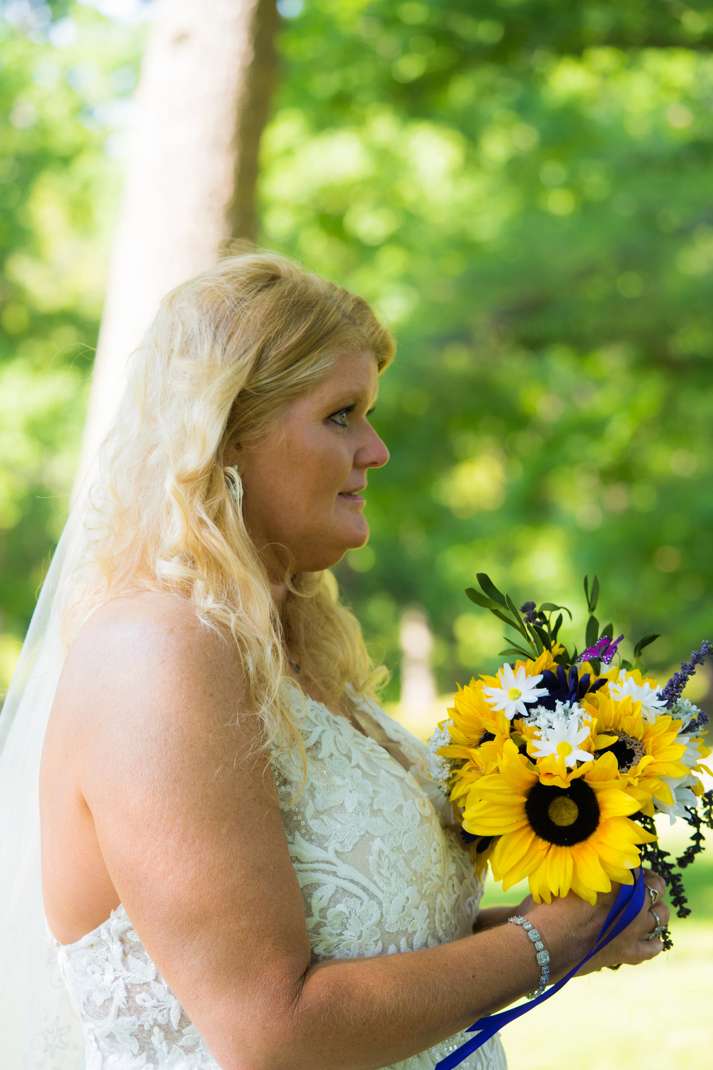 A bride with long blonde hair wearing a white lace dress, holding a bouquet of sunflowers, daisies, and other flowers outdoors on a sunny day.