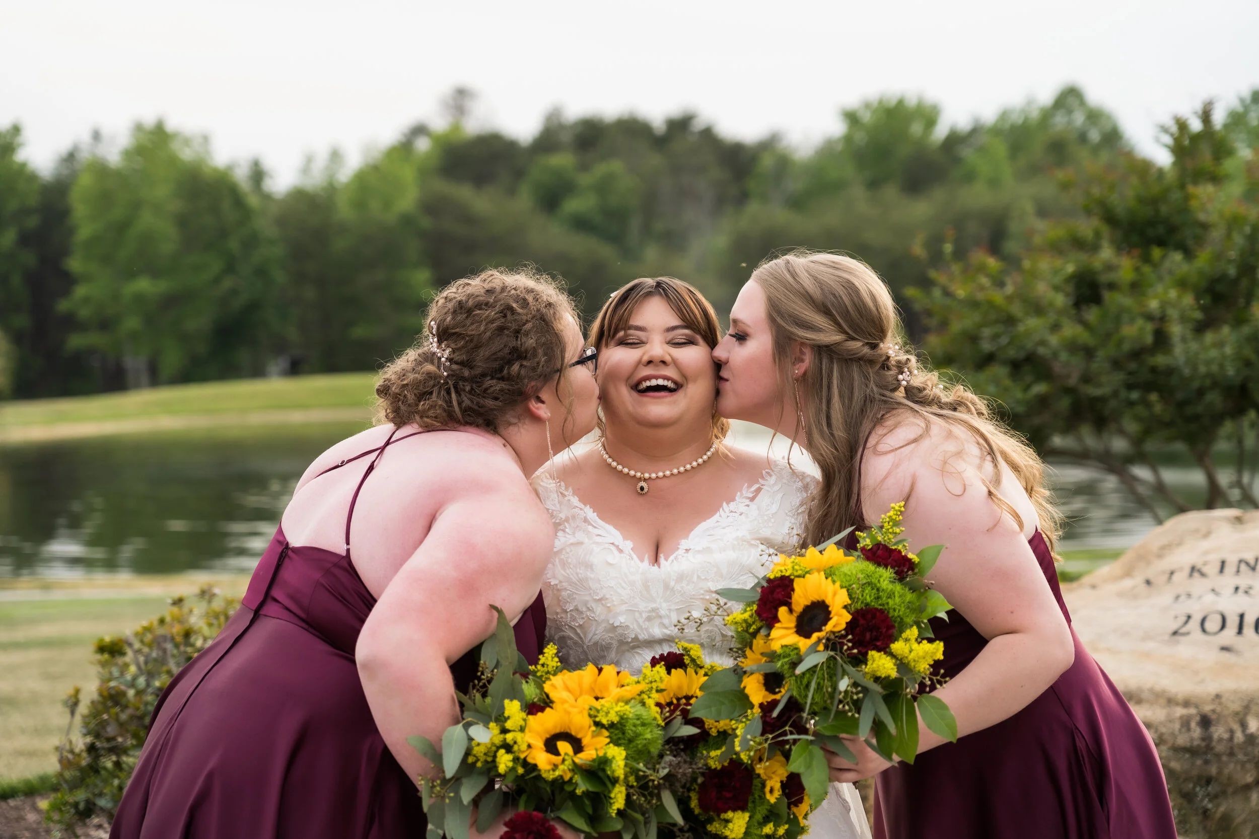 Bride in white wedding dress with two bridesmaids in purple dresses, all smiling, with the bridesmaids kissing the bride on each cheek, holding large bouquets of yellow and burgundy flowers near a lake and trees.
