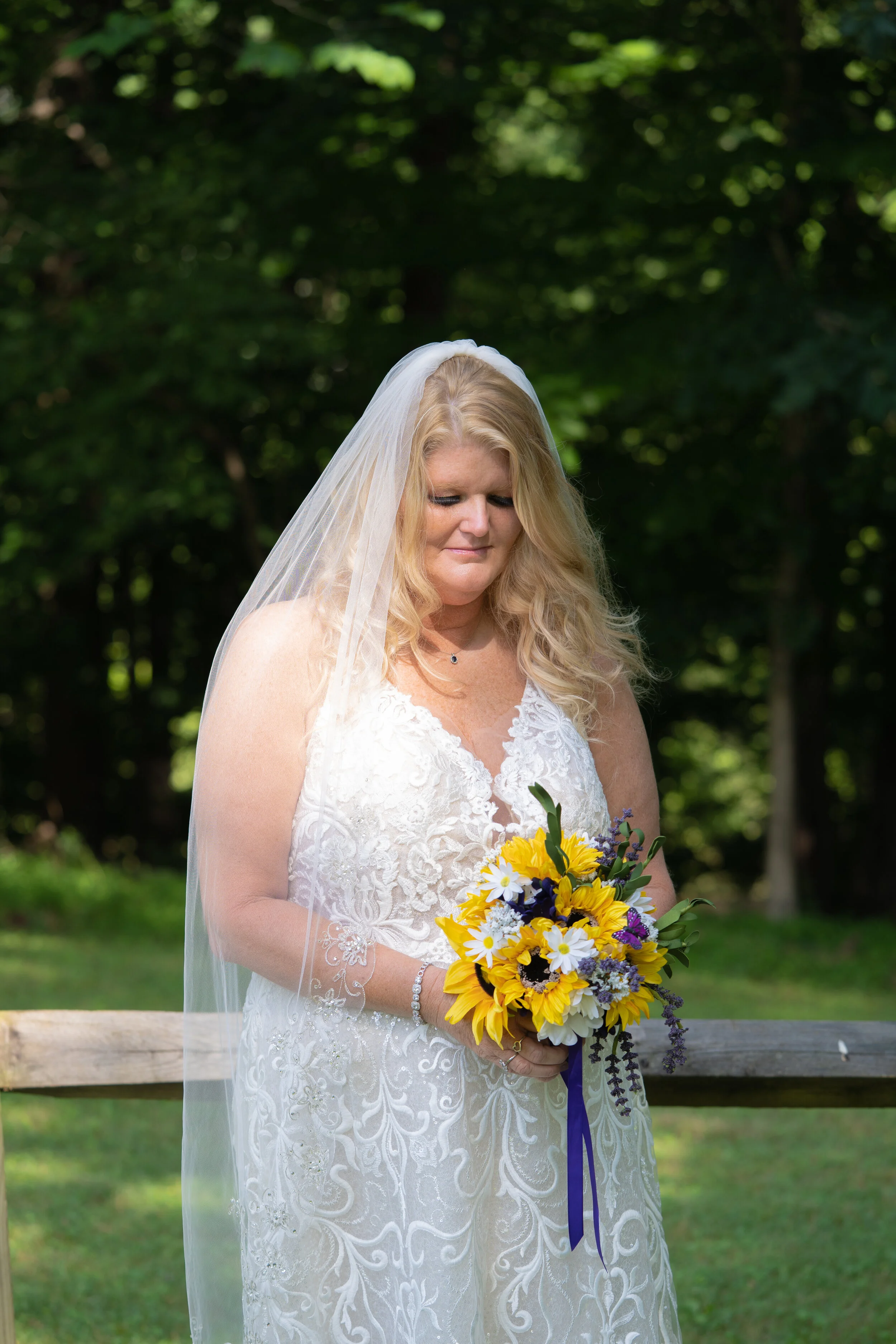 A woman in a white lace wedding dress holding a bouquet of yellow, white, and purple flowers outdoors, with a green tree background.