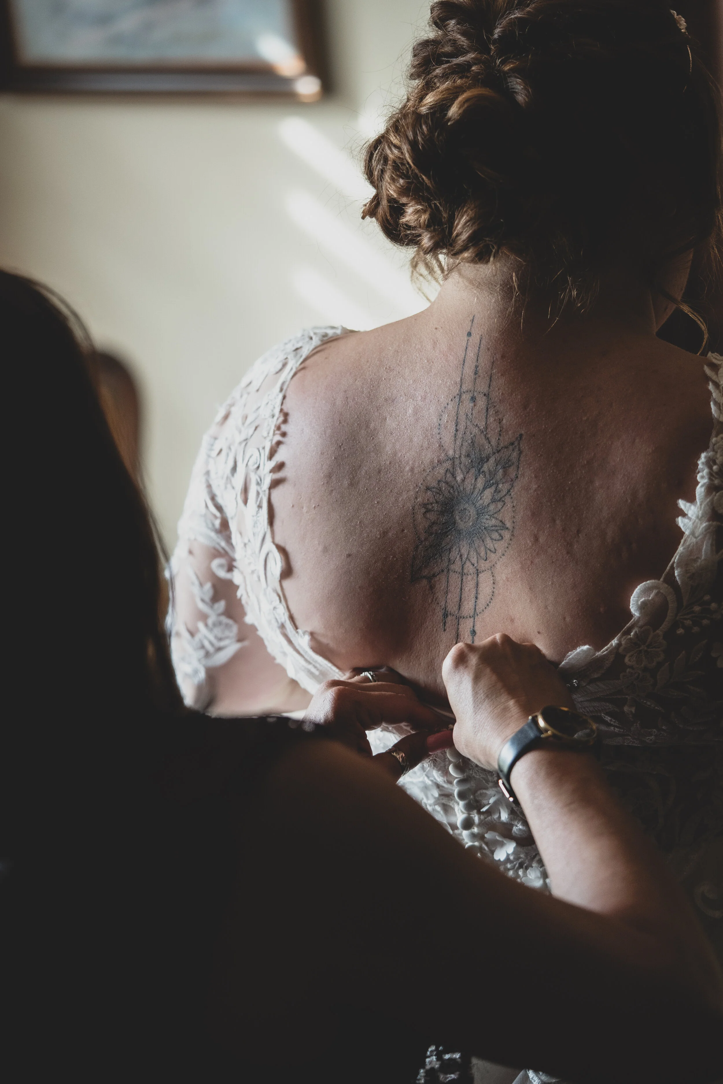 A woman with a floral tattoo on her upper back getting a dress adjusted by another person.