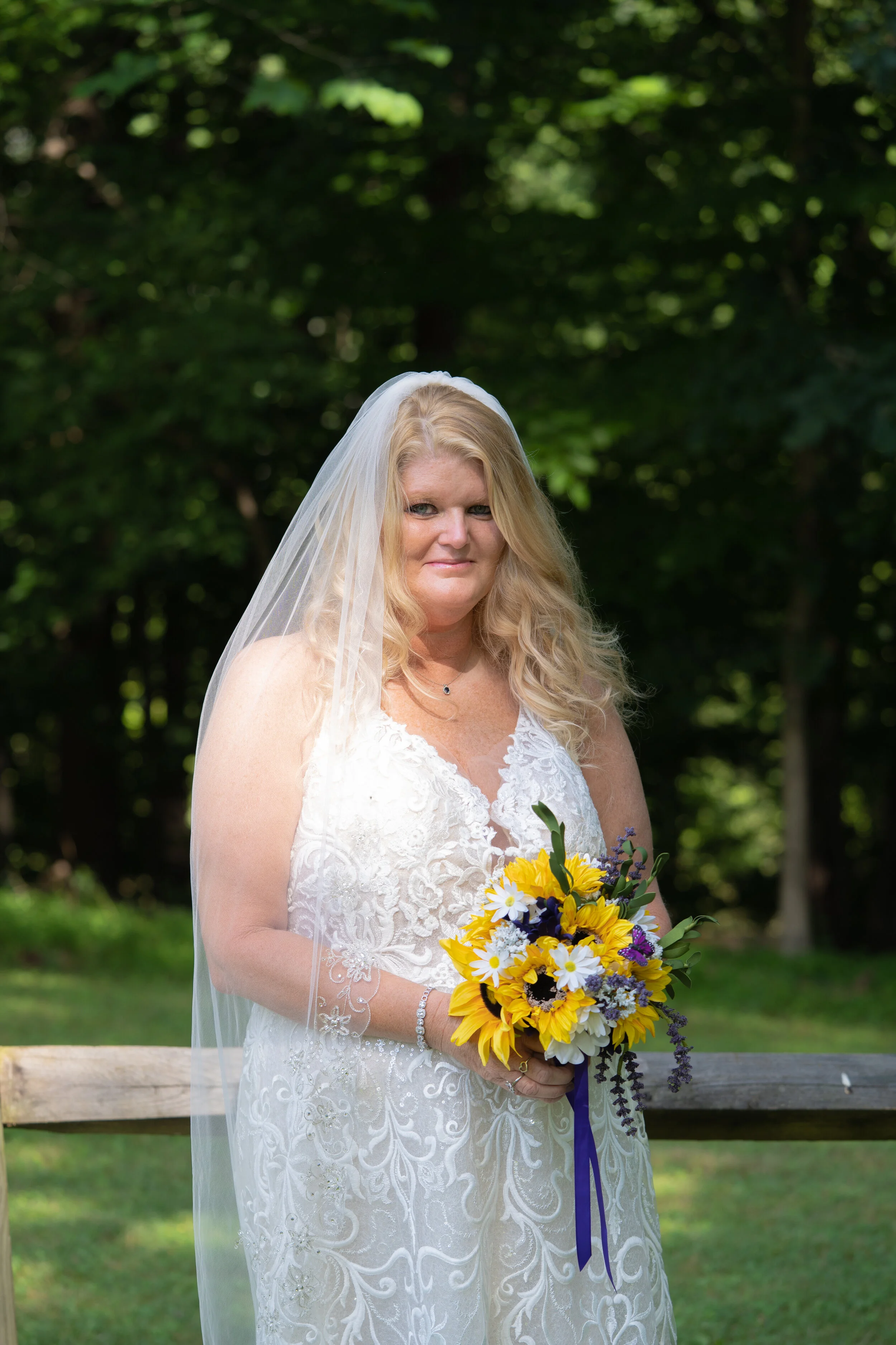A bride with long blonde hair in a white lace wedding dress holding a colorful bouquet of sunflowers, daisies, and purple flowers outdoors during daytime.
