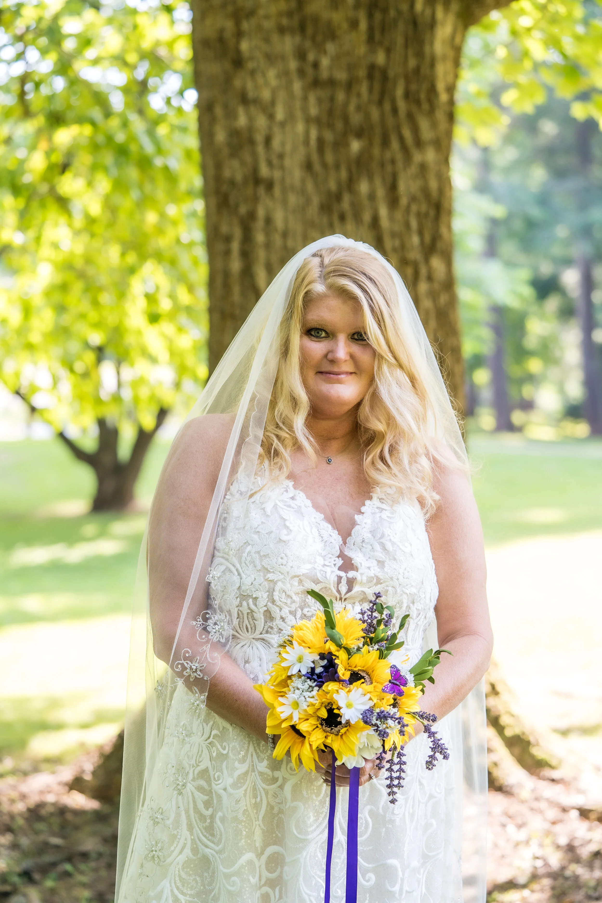 A bride with long blonde hair, wearing a white lace wedding dress and veil, holding a bouquet of sunflowers, daisies, and purple flowers, standing outdoors near a large tree on a sunny day.