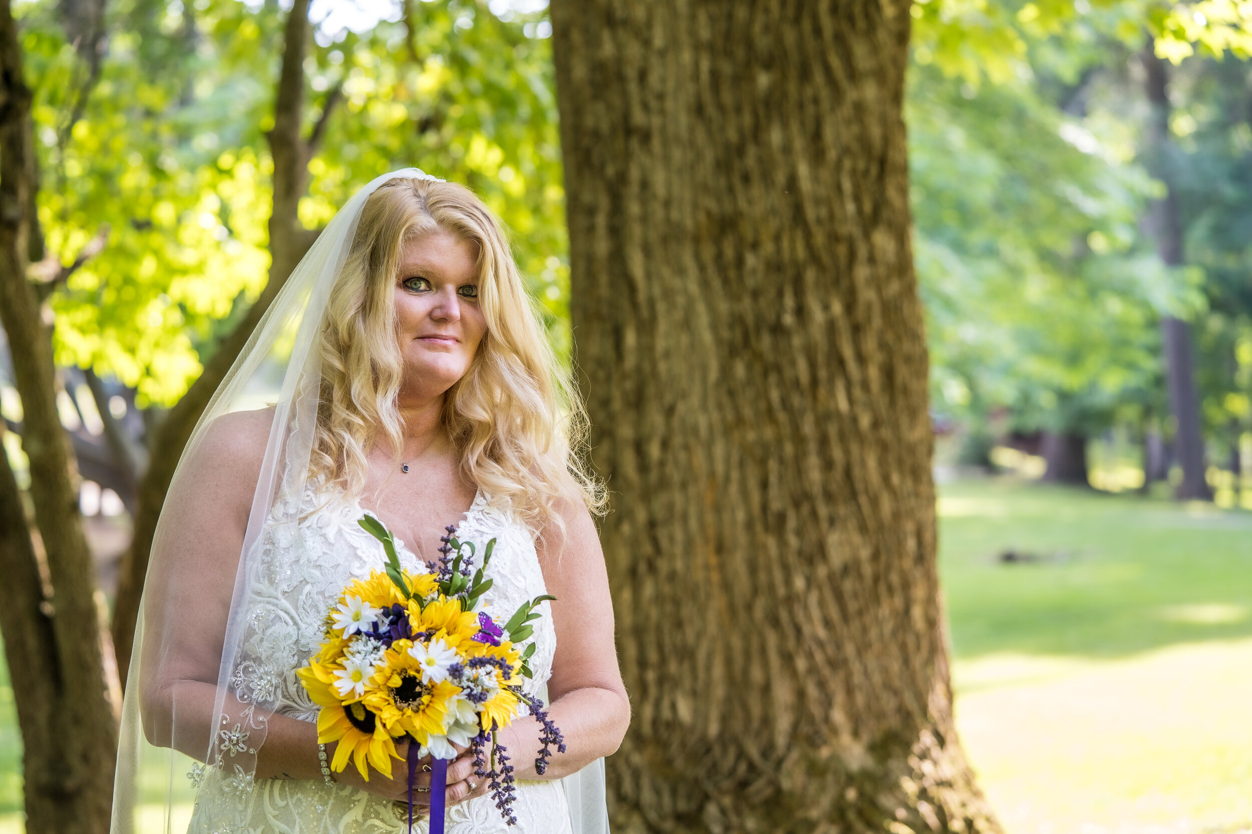 A woman with long blonde hair wearing a white wedding dress and veil, holding a bouquet of yellow, white, and purple flowers, standing outdoors in front of a large tree with green leaves.
