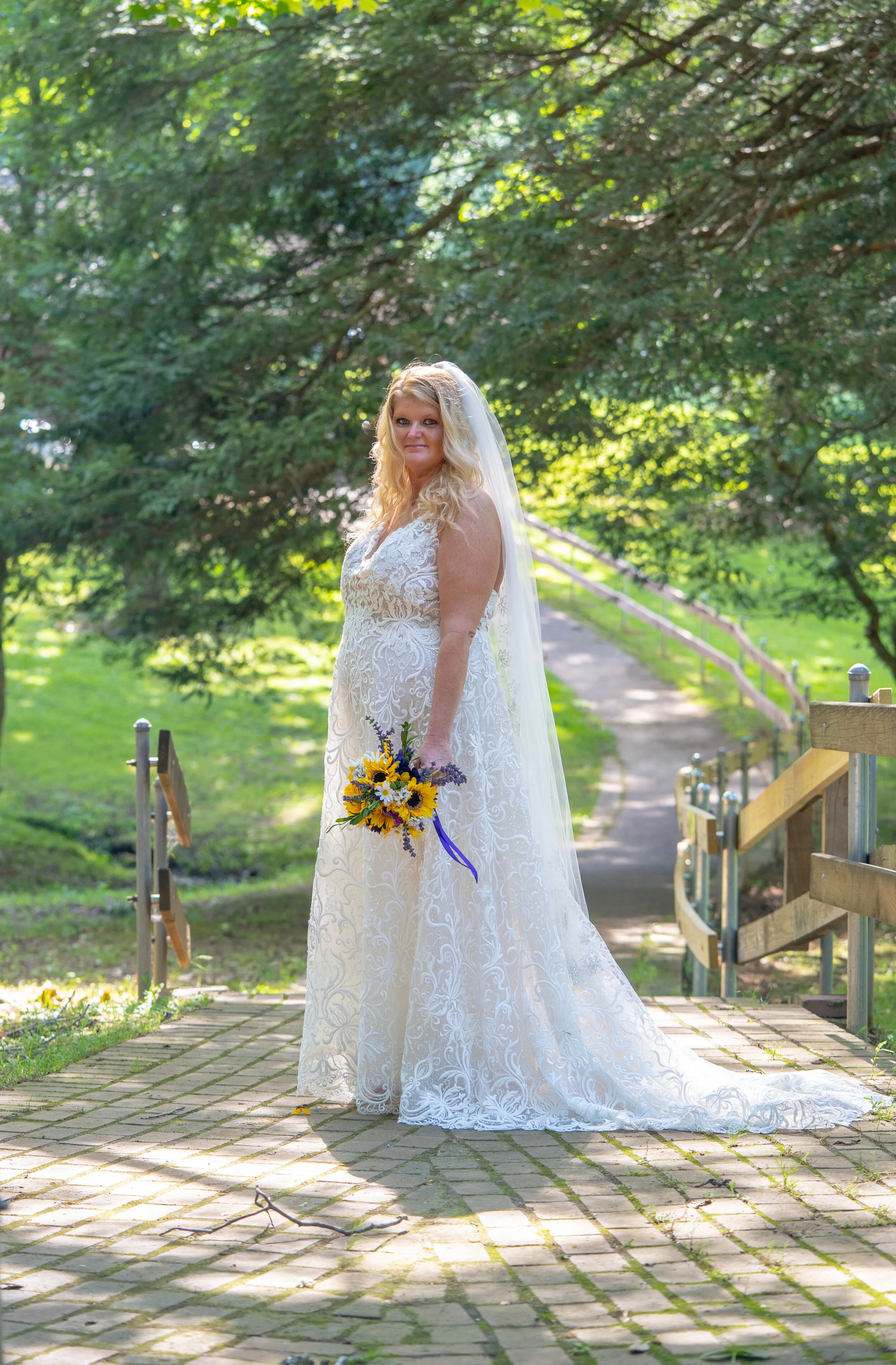 A bride in a white wedding dress holding a bouquet of sunflowers and lavender, standing on a brick pathway in a lush green park with trees.